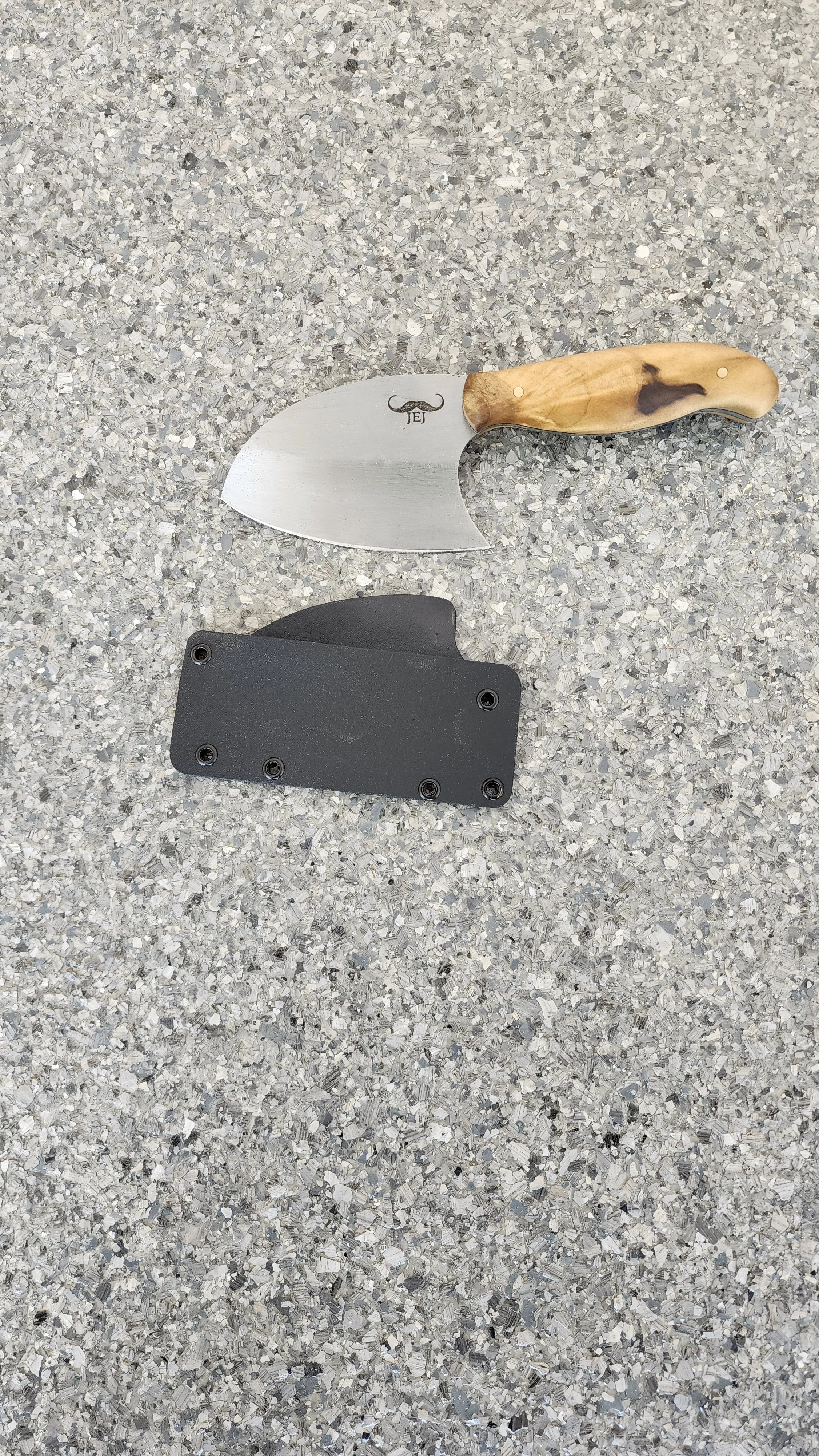 A small curved knife with a wooden handle and a black sheath on a speckled gray surface.