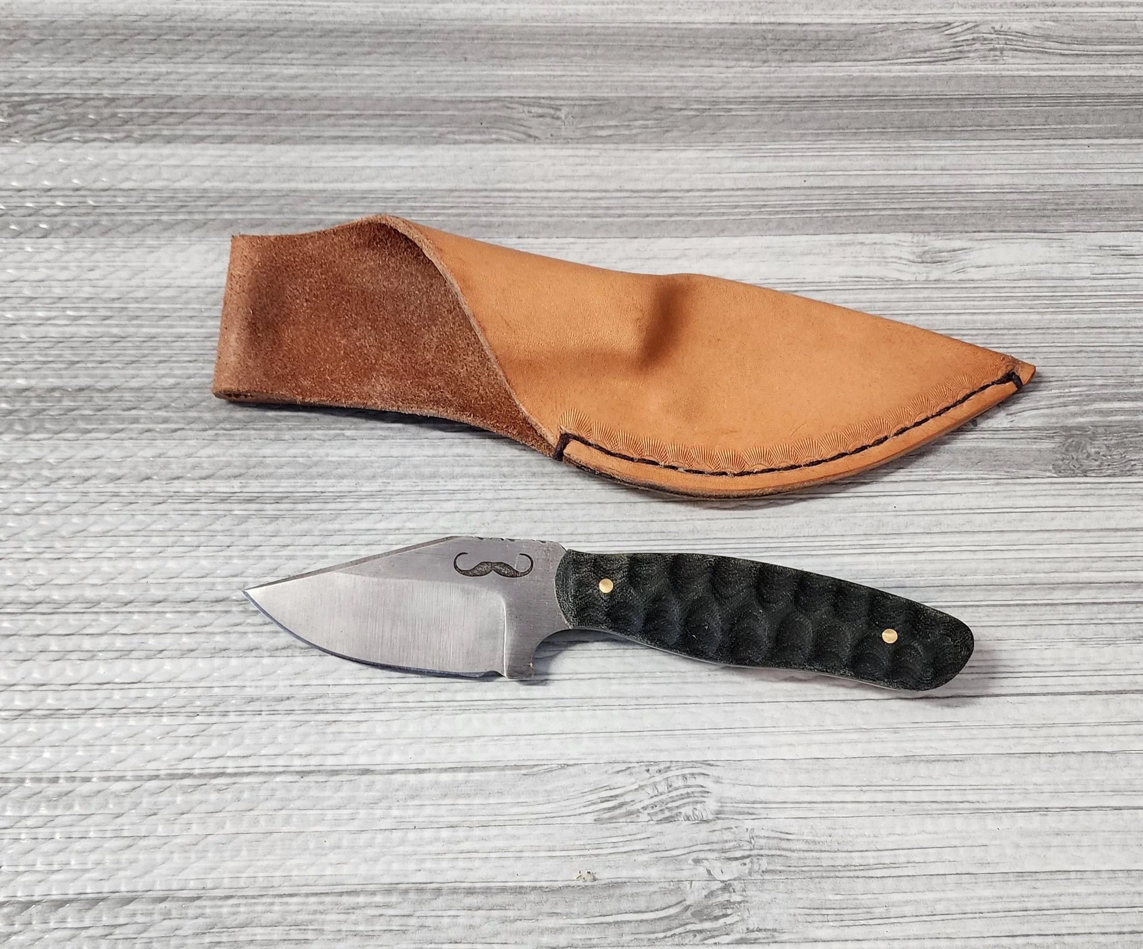 A folding pocket knife with a black textured handle and a curved blade, placed on a light wooden surface next to a leather sheath.