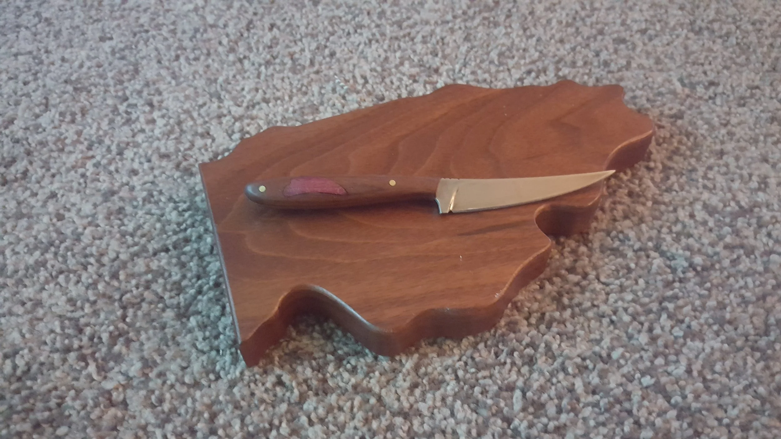Small knife with a wooden handle resting on a wooden leaf-shaped cutting board on a carpeted floor.