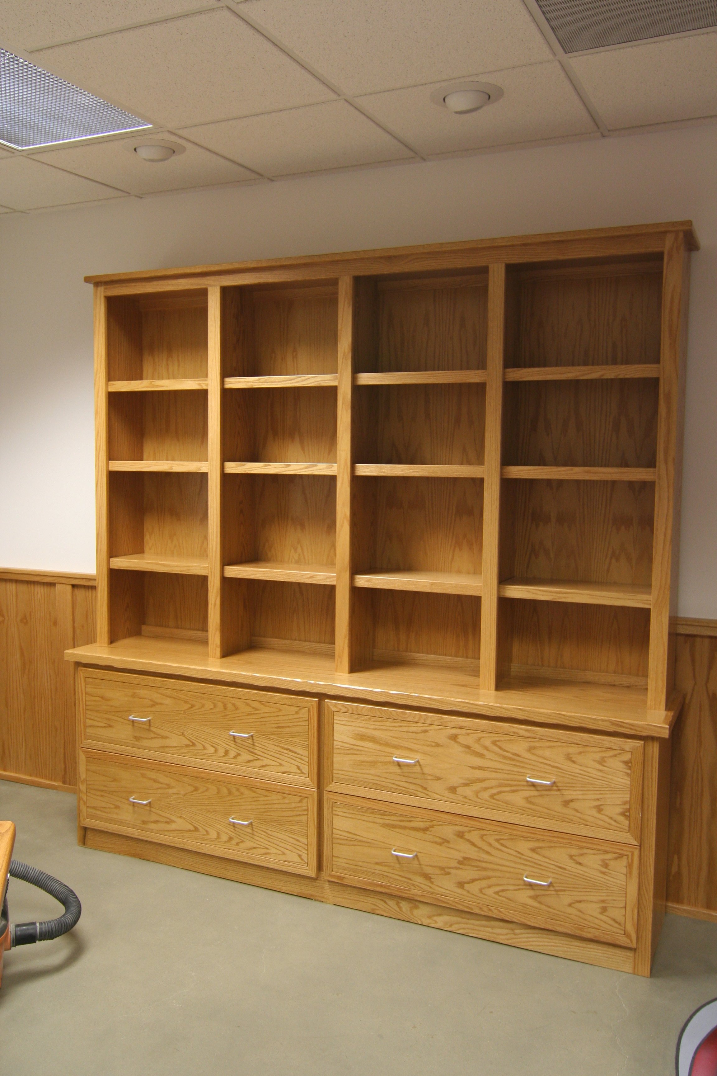 Empty wooden bookshelf with six open shelves and six drawers at the bottom in a room with a drop ceiling.
