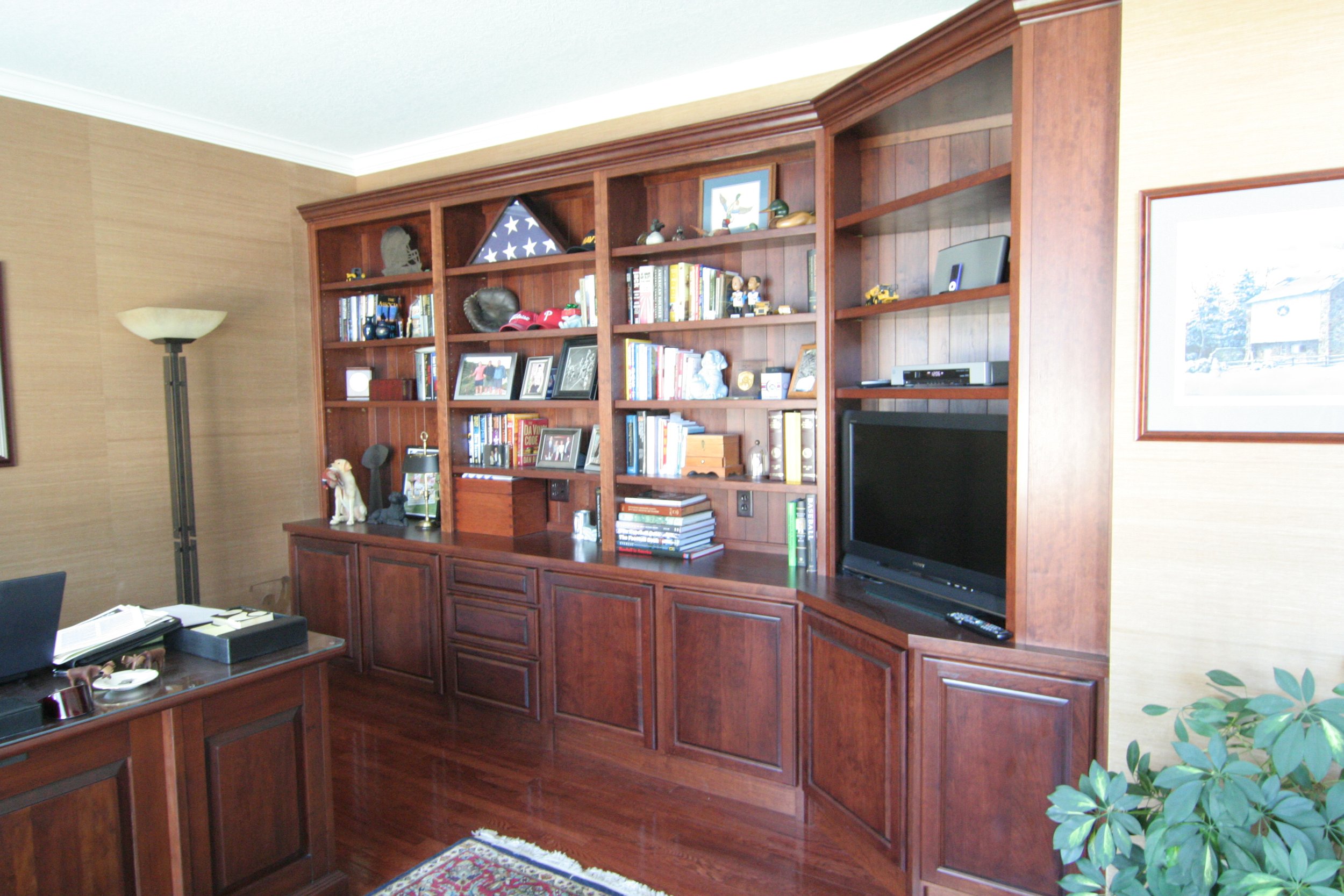 A wooden built-in bookshelf in a living room containing books, photo frames, toys, and decorative items, with a television on the lower right side.
