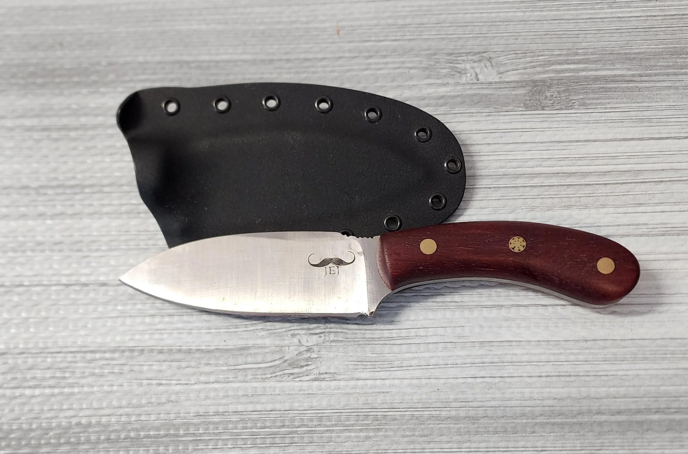 Kitchen knife with a curved red wooden handle and a black protective sheath on a light wood surface.