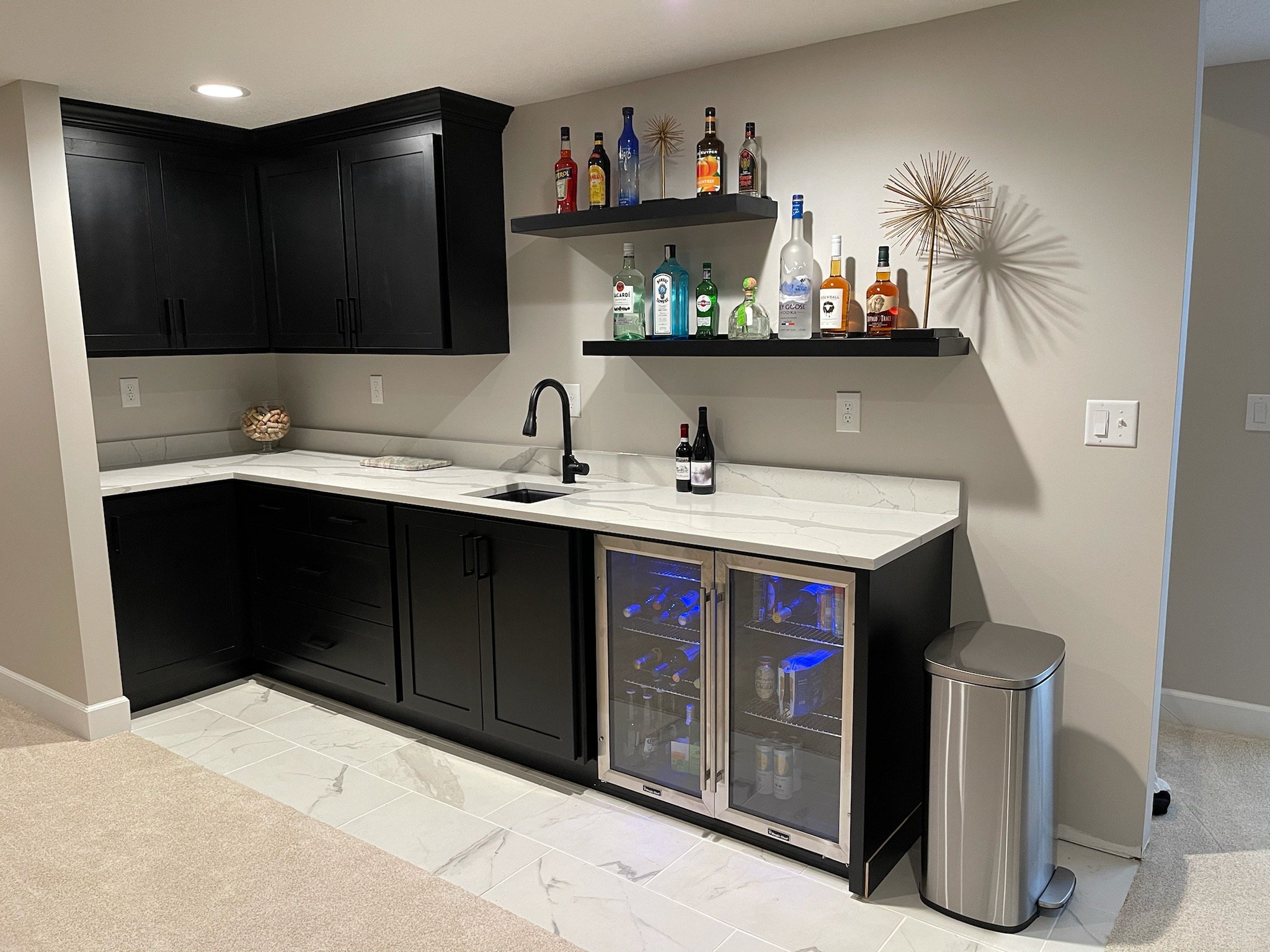 Home bar area with black cabinets, white marble countertop, open shelves with various liquor bottles, a mini wine refrigerator, and a silver trash can.