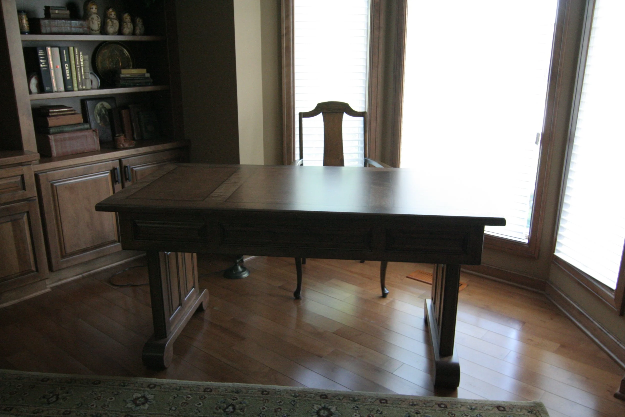 Wooden desk in a corner of a room with wooden floors, a bookshelf on the left, and large windows with blinds on the right.