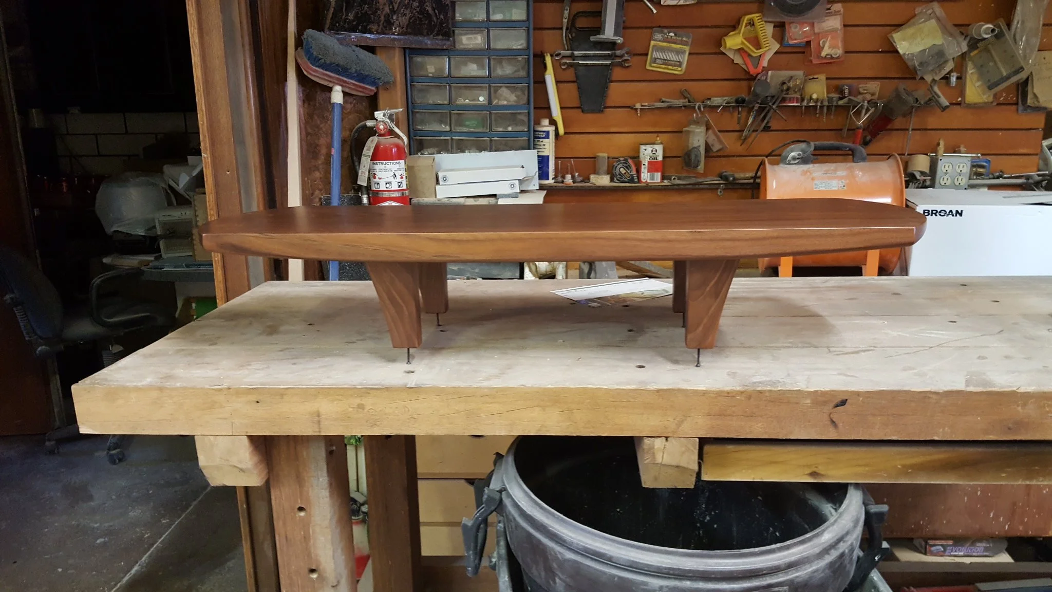 A small wooden table with curved edges and four angled legs, placed on a workbench in a workshop with various tools and supplies on the wall in the background.