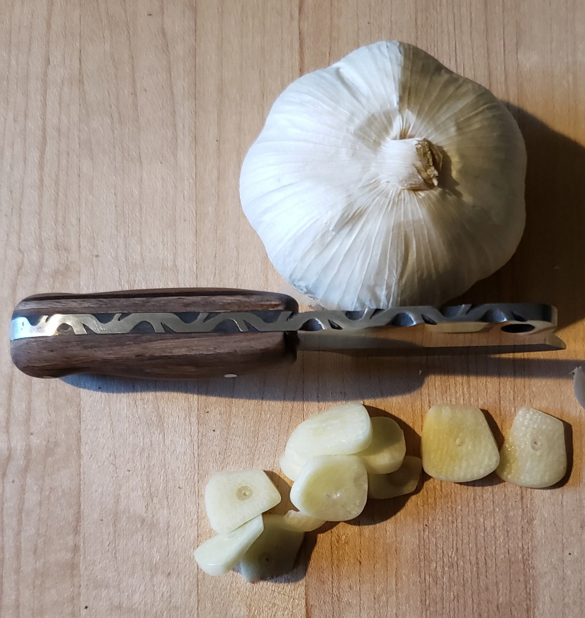 Whole garlic bulb, sliced garlic cloves, and a garlic knife on a wooden cutting board.