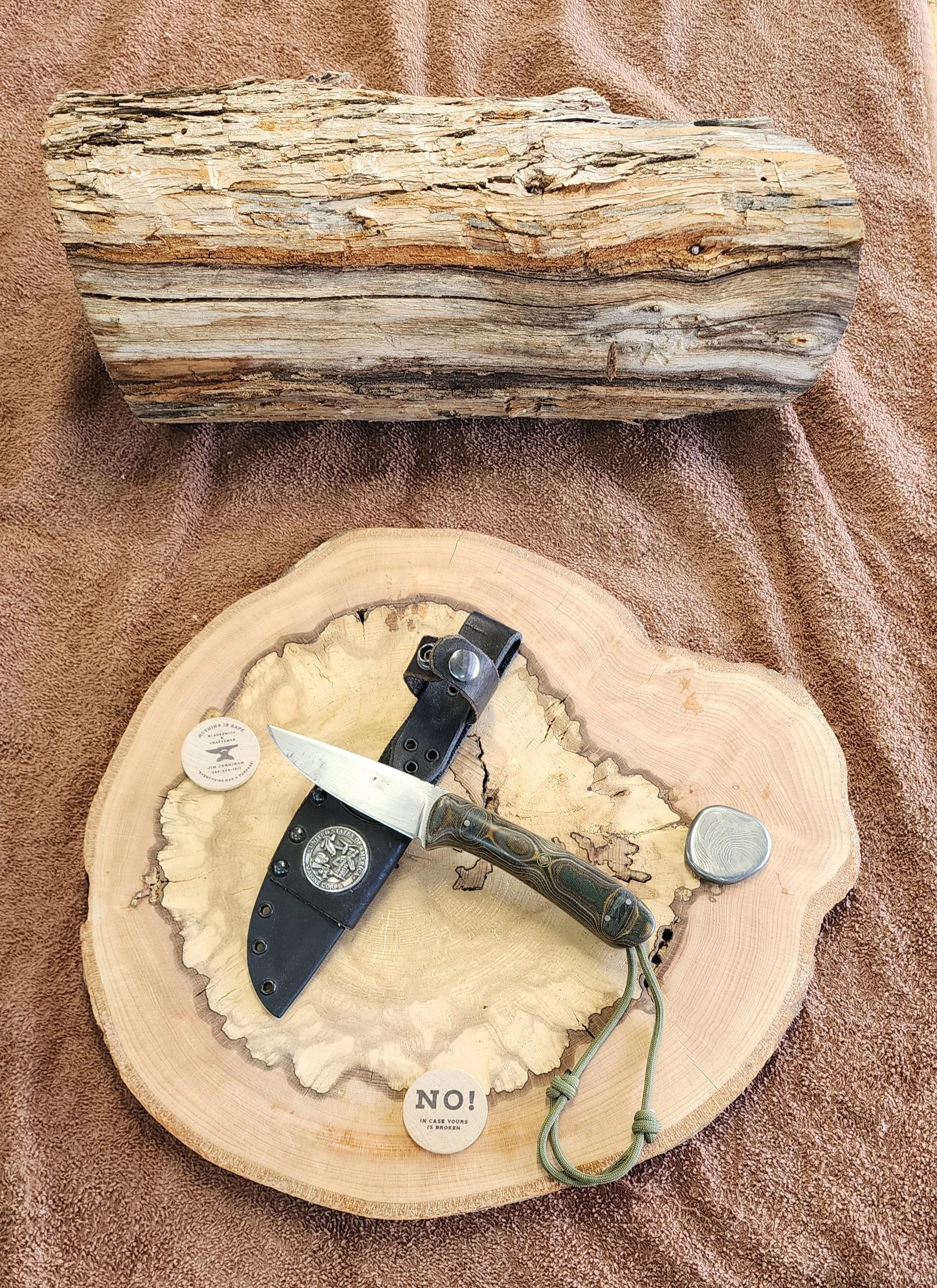 A wooden log at the top and a cross-section of a tree trunk with a knife, a small container, and a button or badge on it, placed on a brown fabric surface.