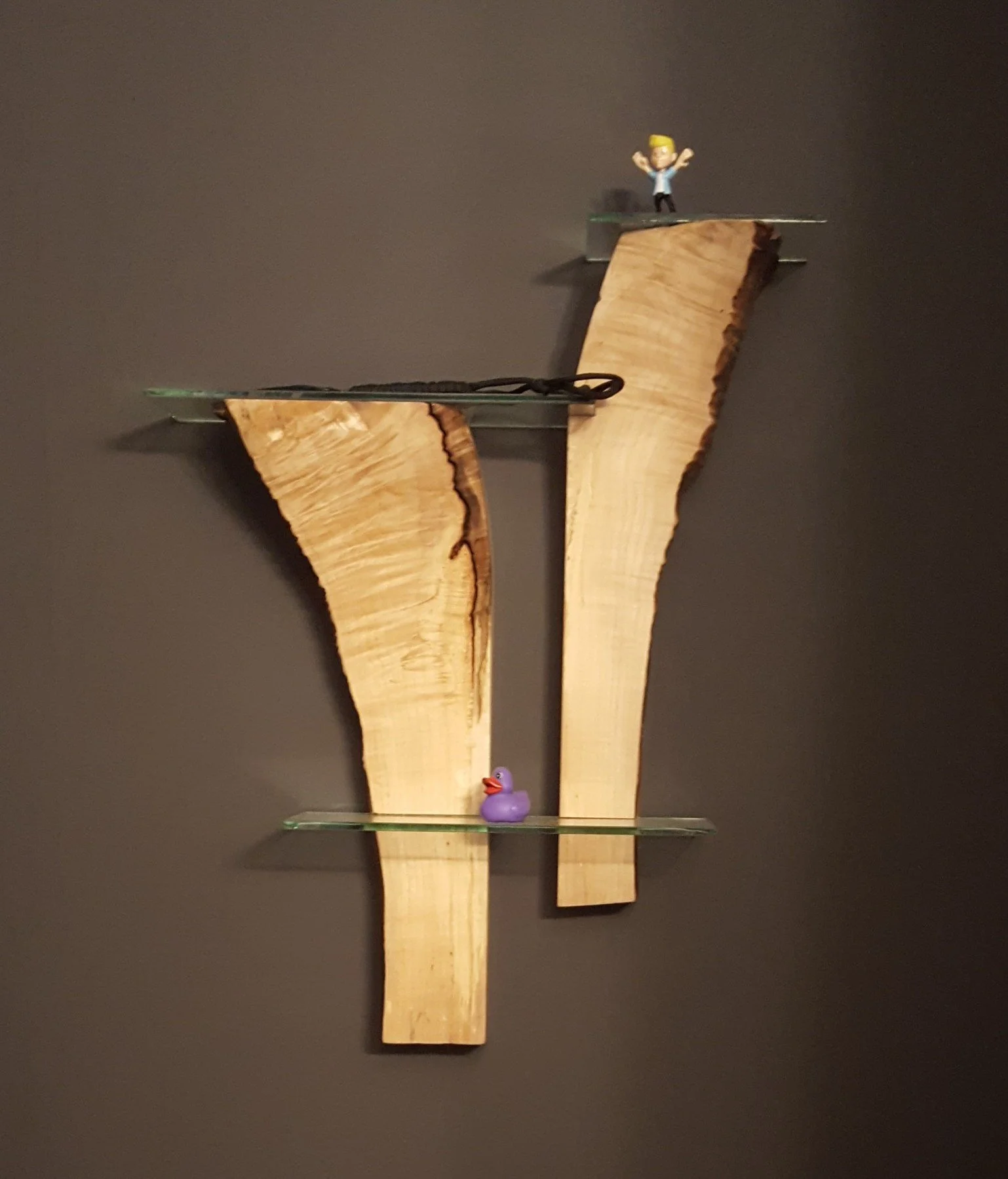 Decorative wall shelf made of raw wood pieces with glass shelves, holding a small rubber duck, a tiny person figurine, and an extra small figurine of a person.