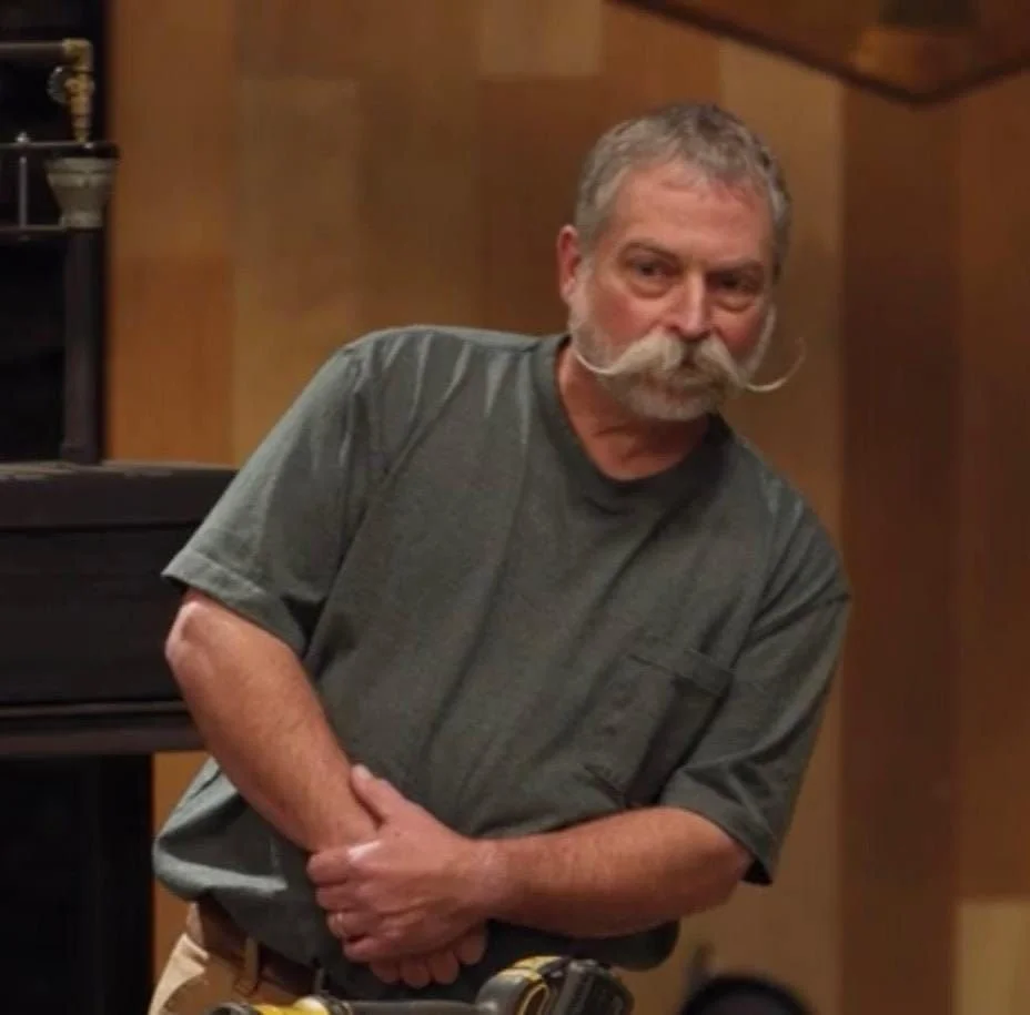 A man with gray hair and a mustache, wearing a dark gray T-shirt, standing with his arms crossed in a room with wooden walls and furniture.