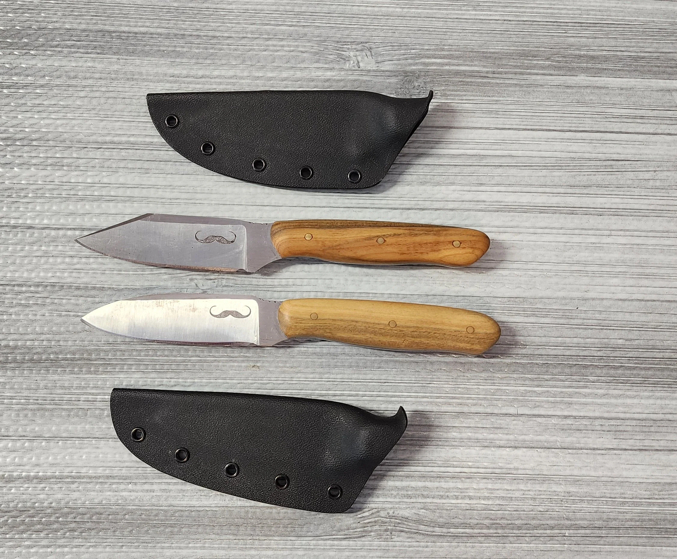 Two fixed-blade knives with wooden handles and decorative mustache engravings on the blades, each paired with a black sheath, are laid out on a gray wooden surface.