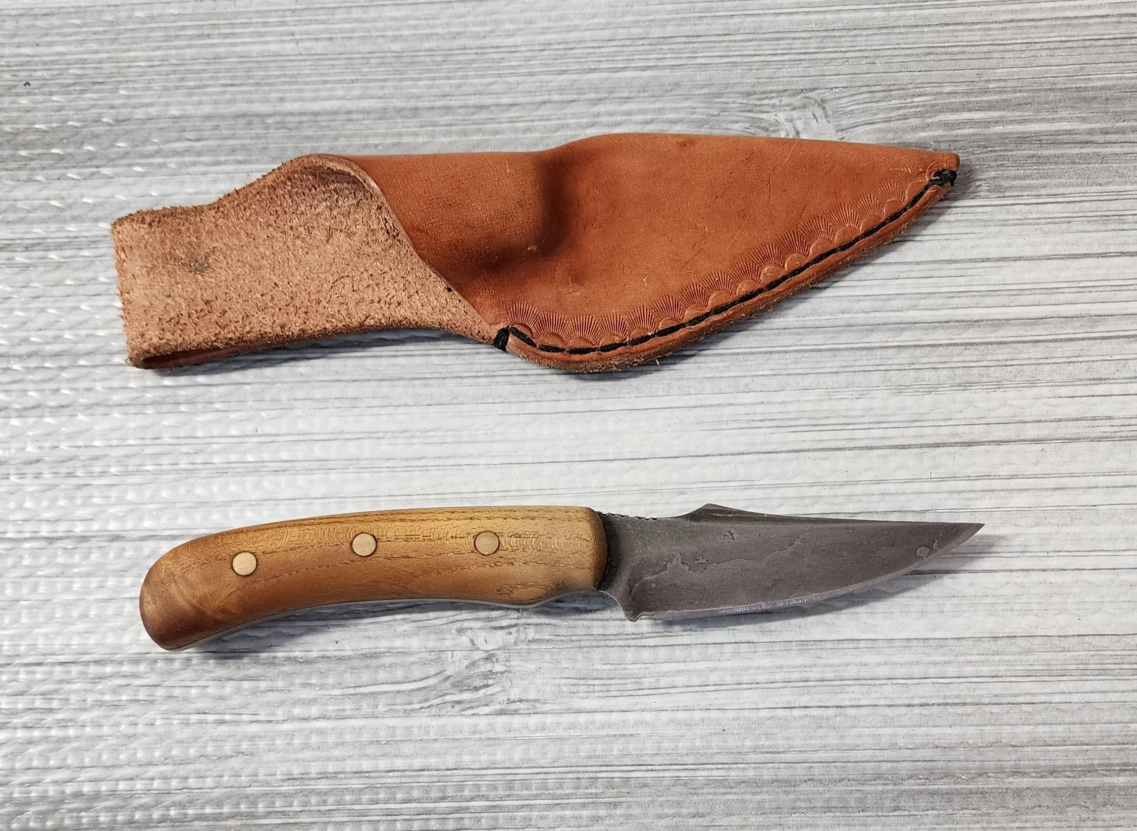 A small hunting or utility knife with a stainless steel blade and a wooden handle, accompanied by a brown leather sheath with decorative stitching.