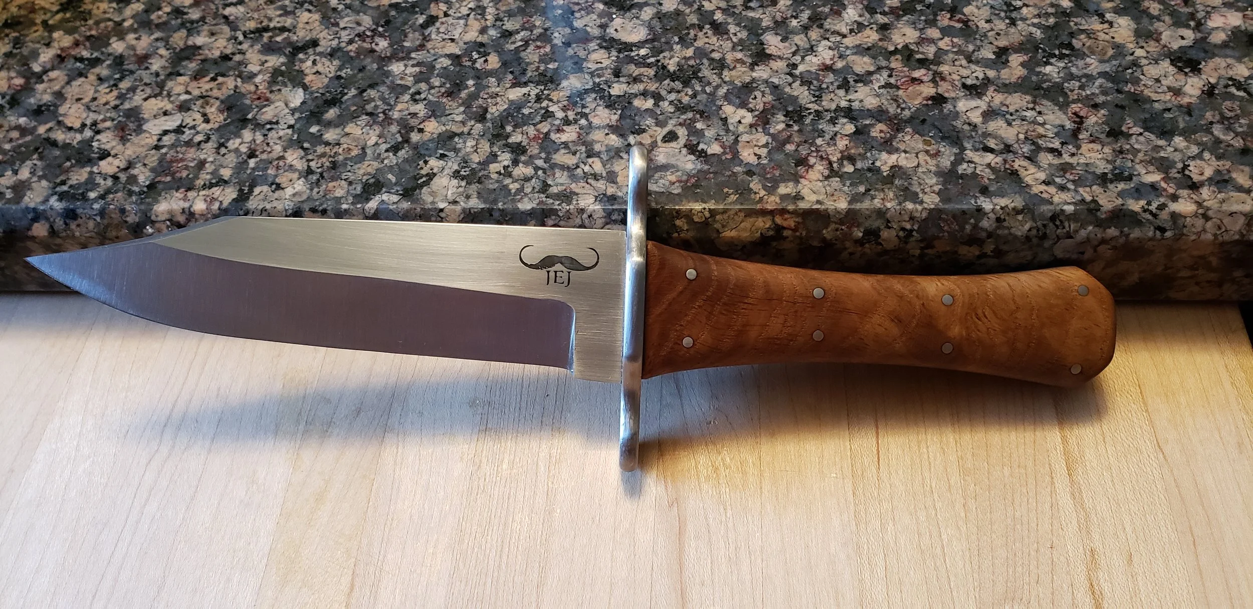 Kitchen knife with a wooden handle, metal guard, and a blade with a mustache logo, resting on a light wooden surface in front of a dark granite countertop.