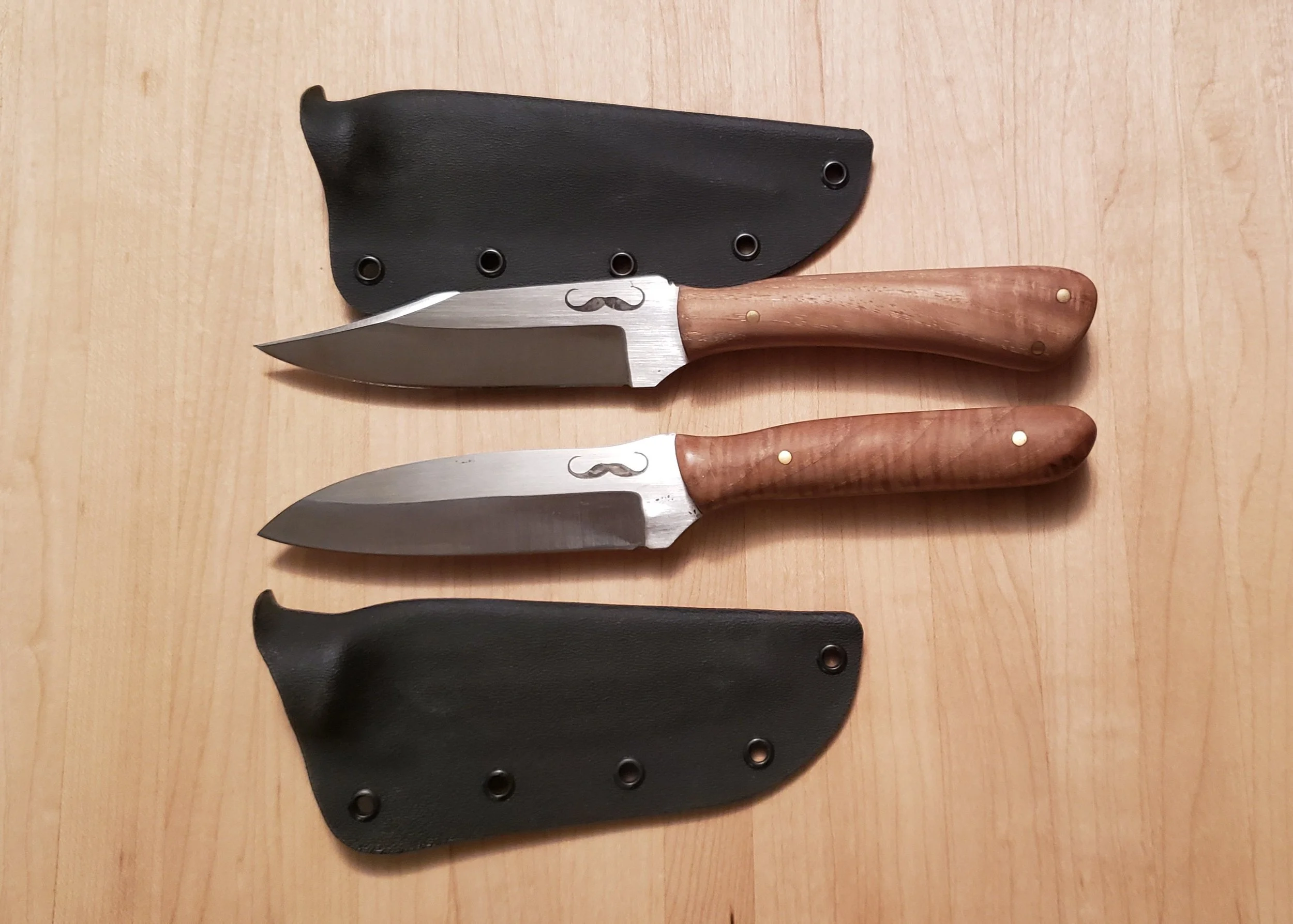 Two fixed-blade knives with wooden handles and brass pins, each with a black holster, on a wooden surface.