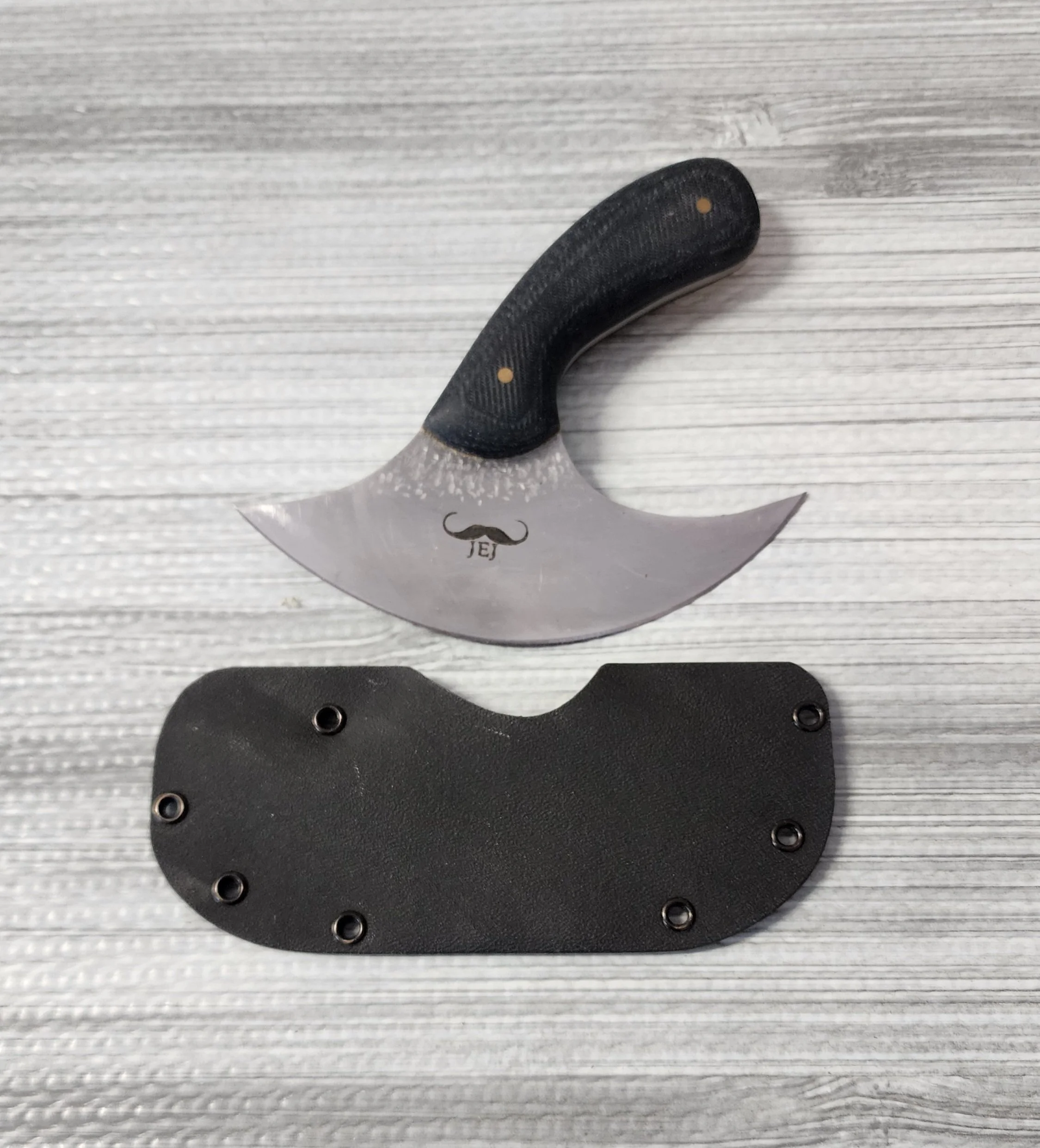 A curved knife with a black handle and a mustache logo on the blade, placed above a black sheath on a wooden surface.