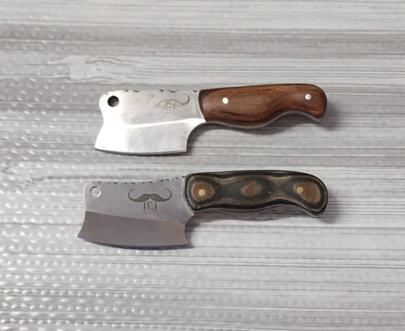 Two small knives with curved blades and wooden handles, one with a plain wooden handle and the other with a black and brown textured handle, placed on a gray textured surface.