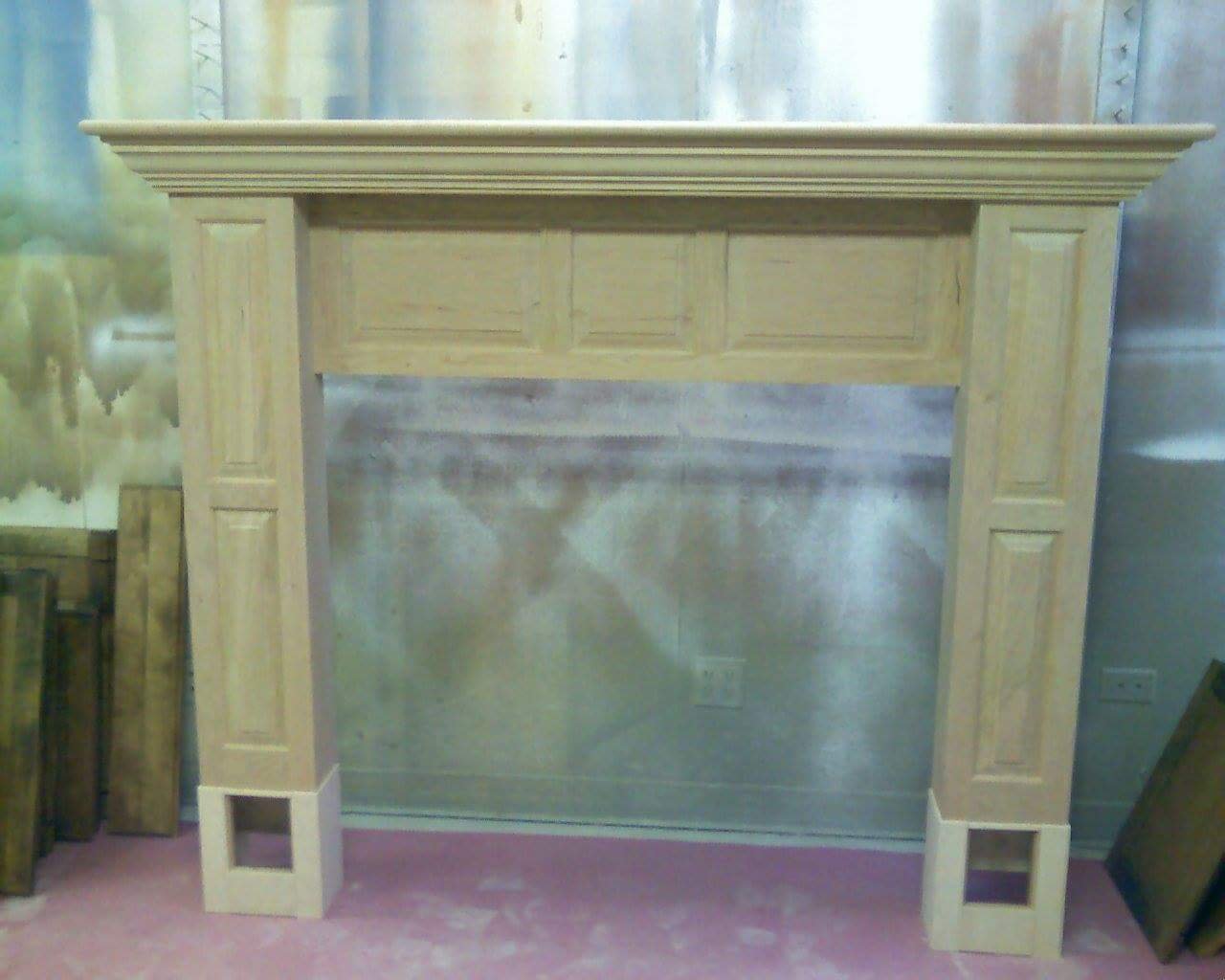 Unfinished wooden fireplace mantel with decorative molding, standing in a workshop with tools and wood pieces nearby.