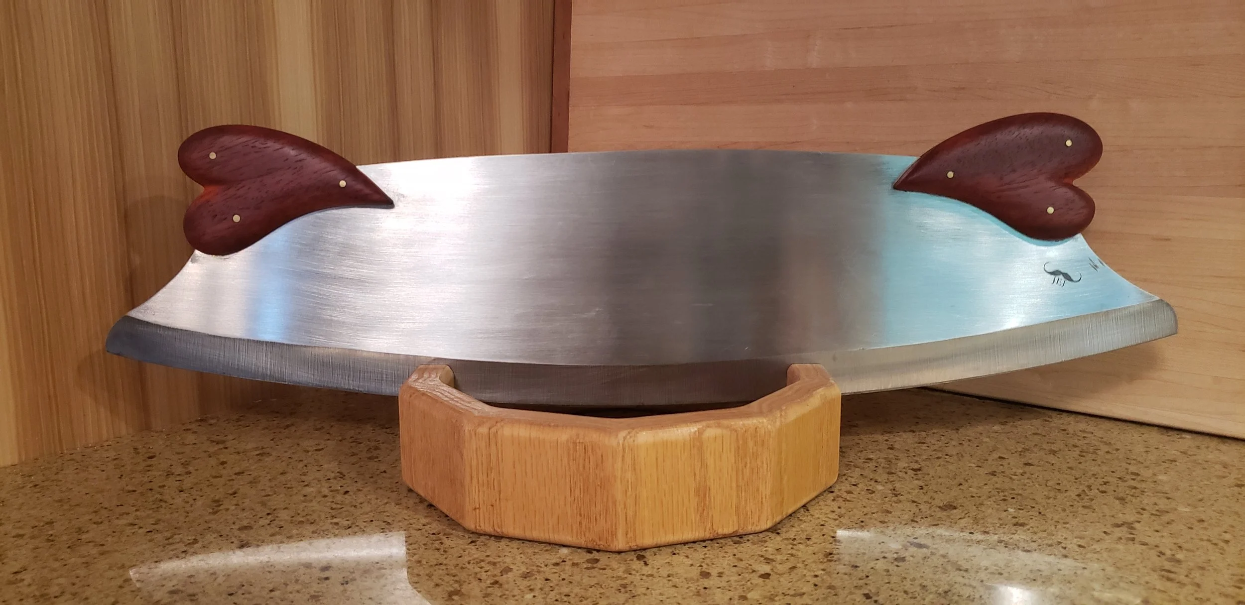 A large meat cleaver with a stainless steel blade and dark wooden handle rests on a round wooden stand on a kitchen countertop. The handle has two brass rivets, and there is a small logo of a mustache near the edge of the blade.
