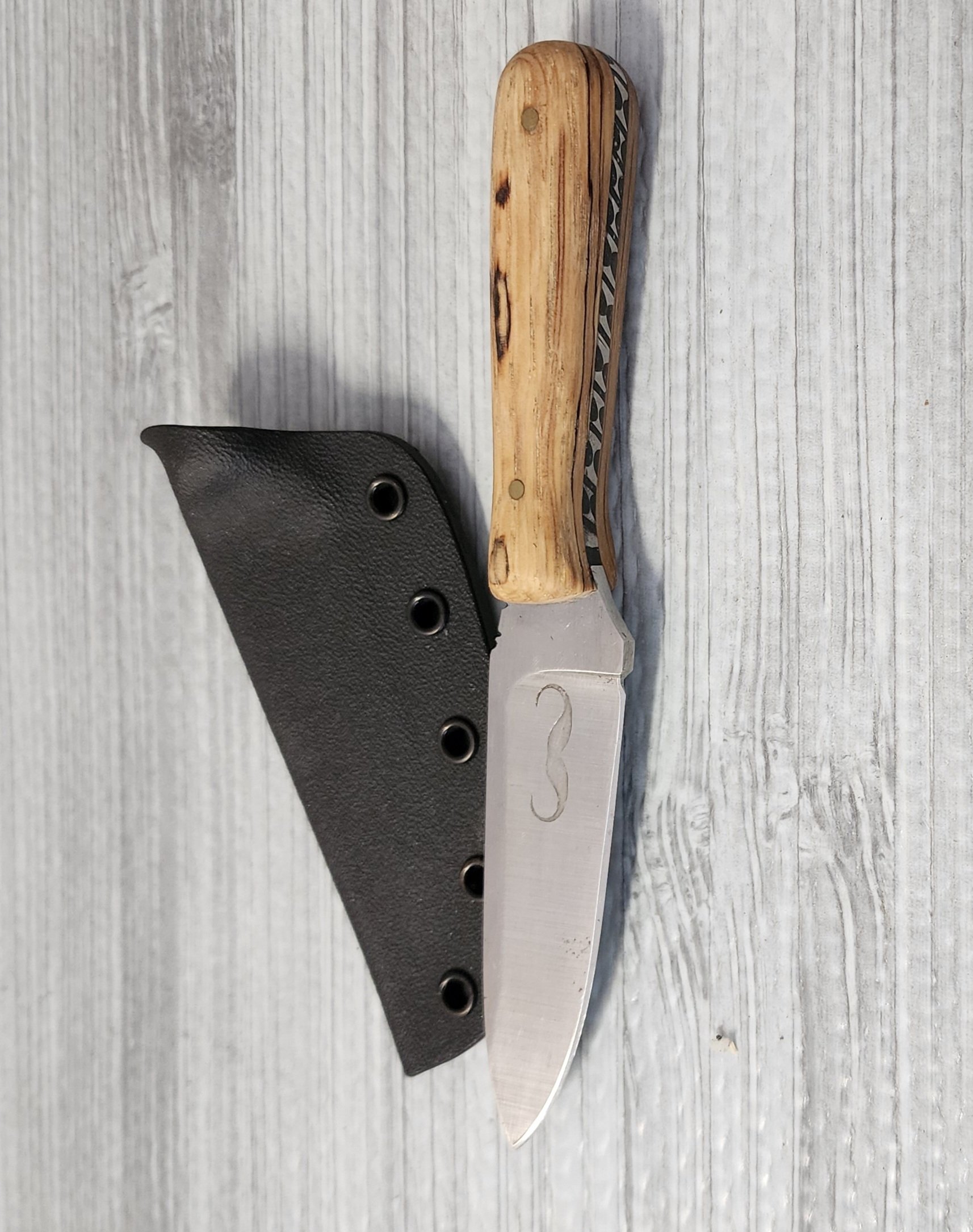 A fixed-blade knife with a wooden handle and a etched logo on the blade placed next to a black sheath on a light gray wooden surface.