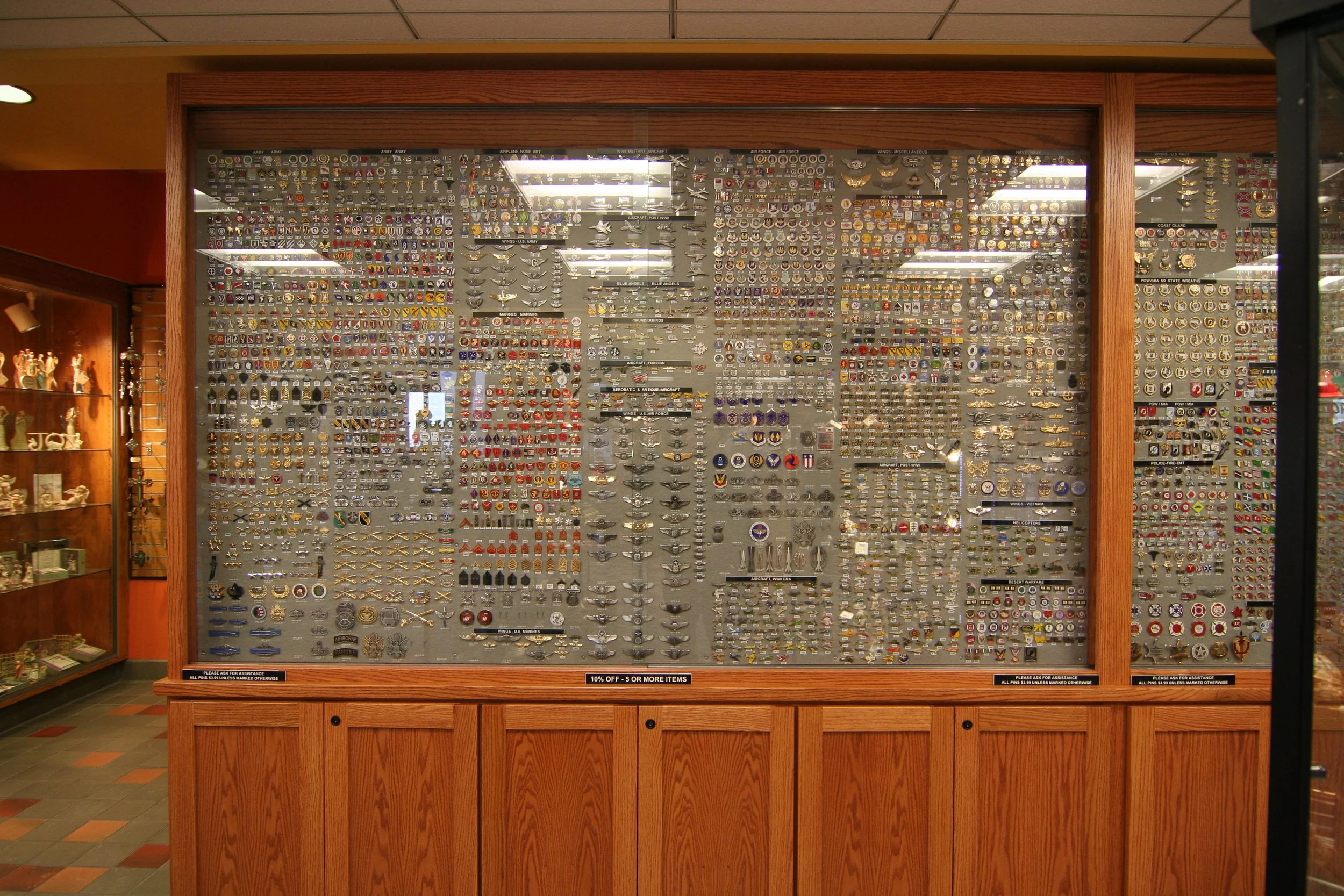 A display case filled with numerous military and aviation badges, patches, and medals. The case is made of wood with a glass front, located inside a store, with shelves of merchandise on the right and a showroom on the left.
