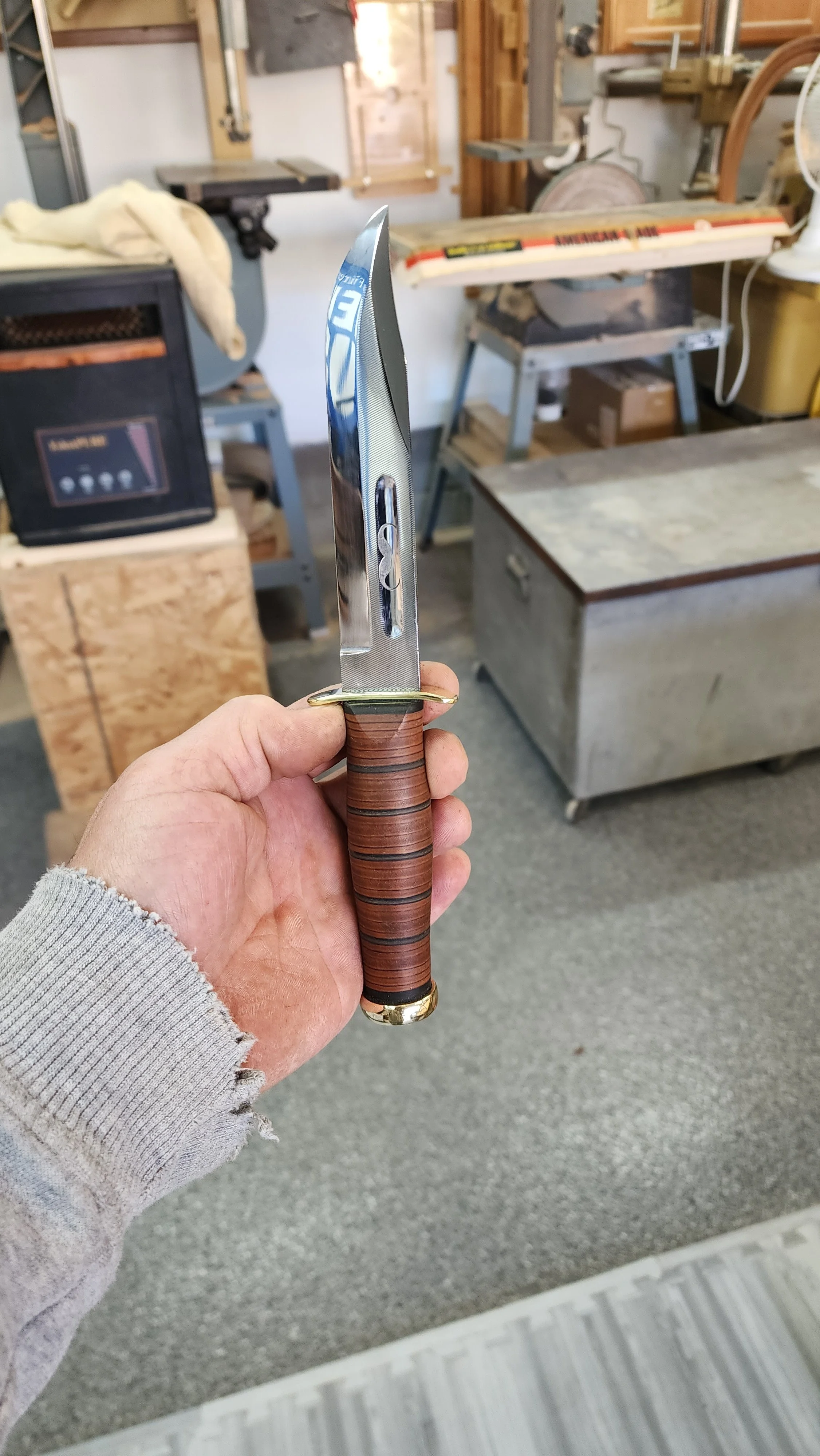 A person holding a dagger with a stainless steel blade and a wooden handle in a workshop setting.