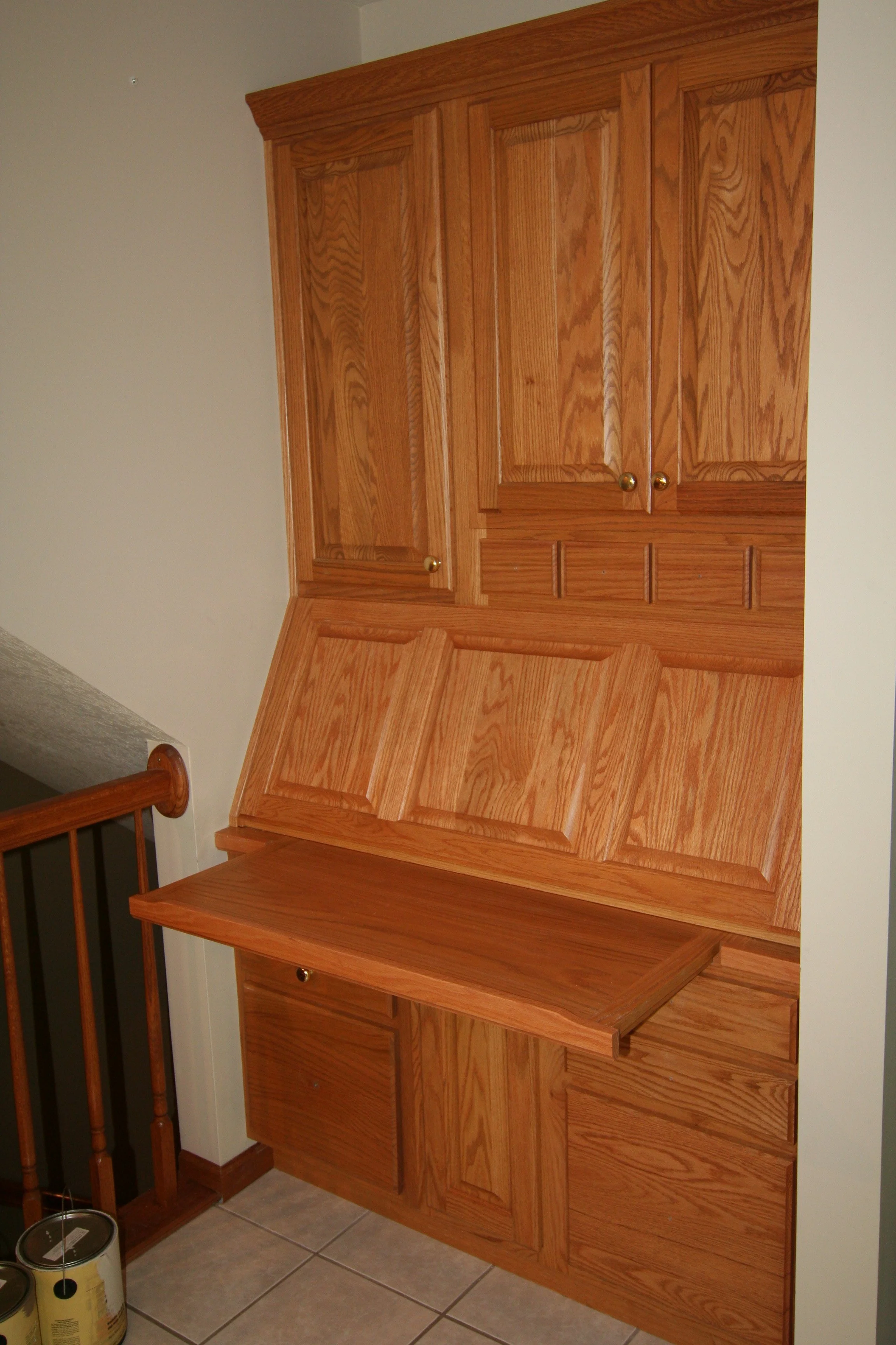 A large wooden built-in wall cabinet with upper and lower storage sections, situated in a room with tiled flooring and a staircase nearby.