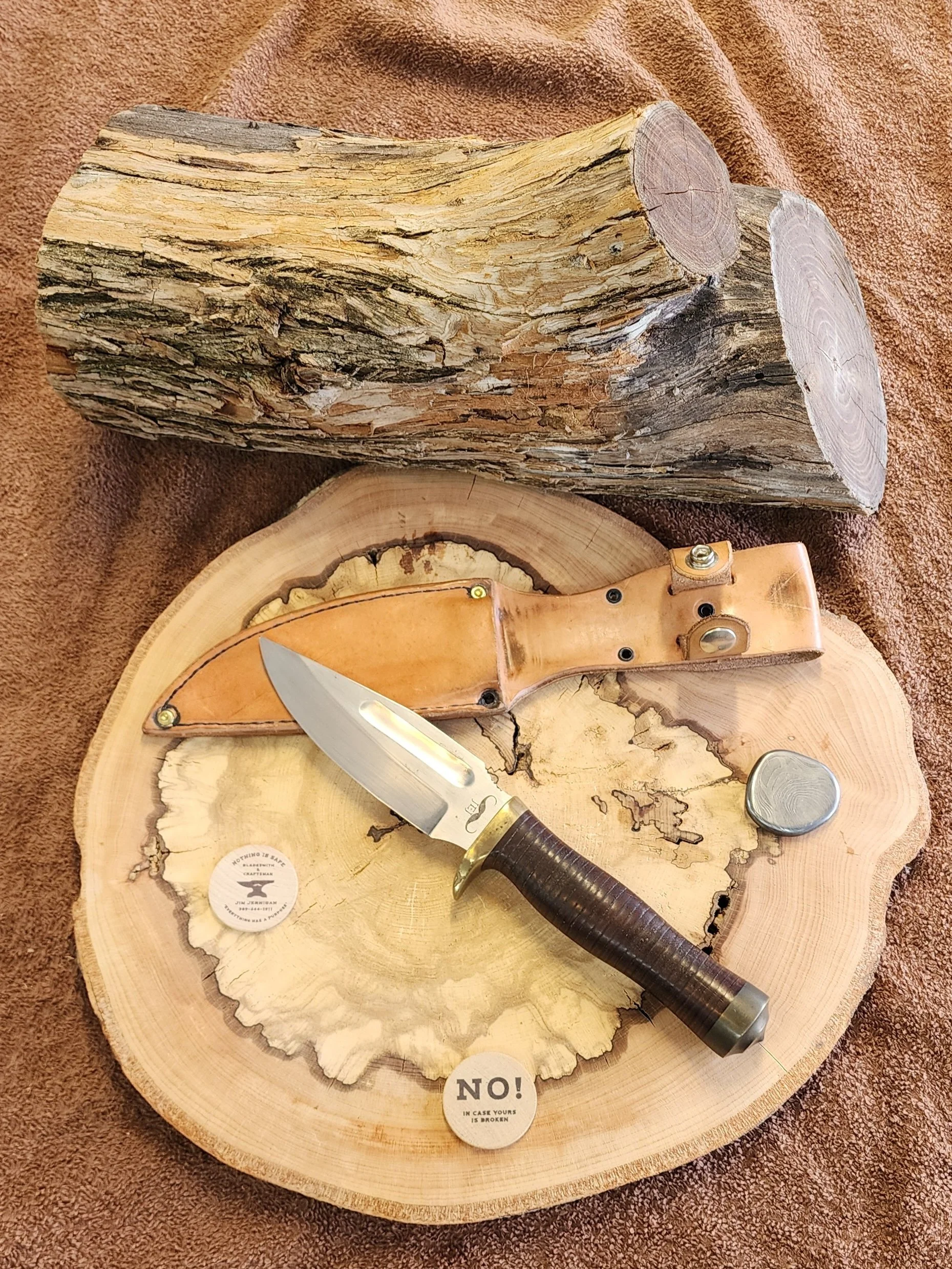 A large cut of wood, a fixed-blade knife with a dark handle, a leather sheath, a small grey stone, and a small round sticker with text are arranged on a cross-section of a tree trunk, with a large log in the background on a brown fabric surface.