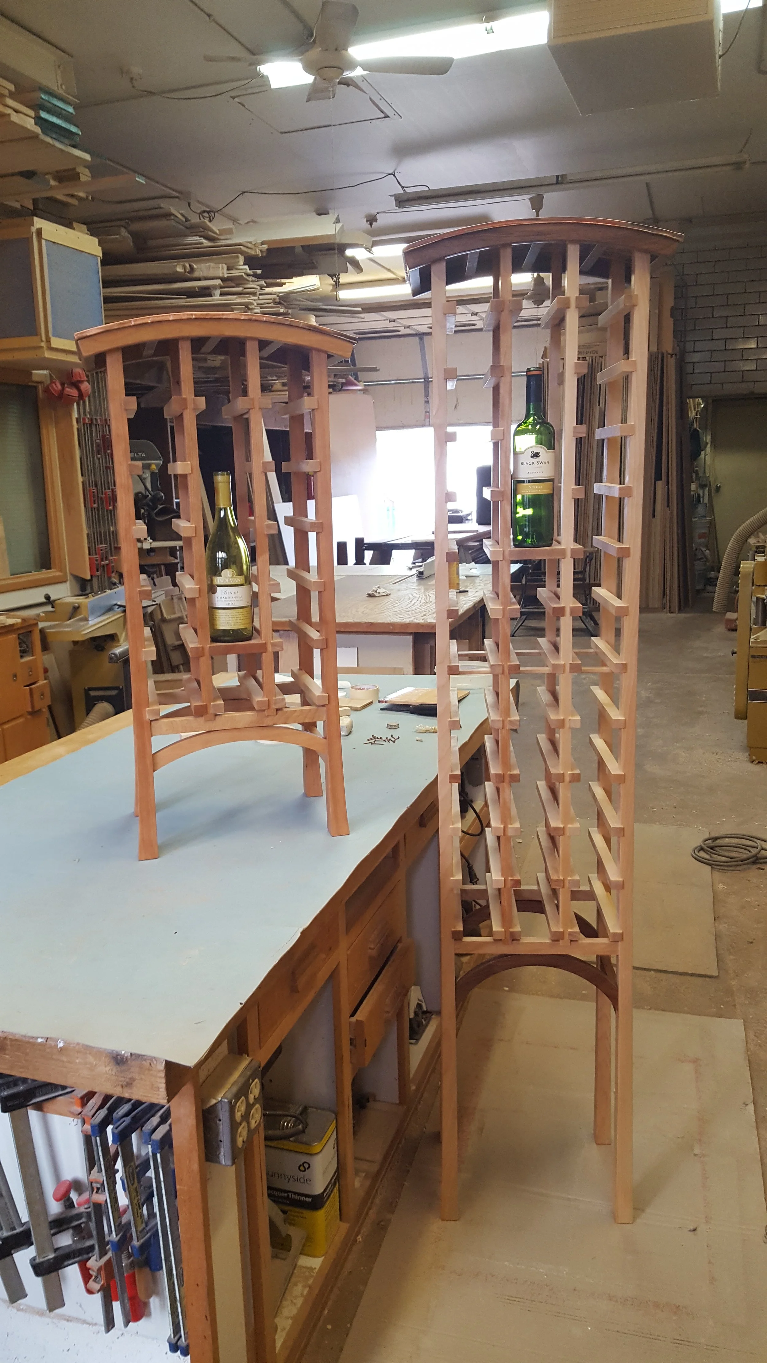 Two wooden wine racks on workbenches in a woodworking shop, each holding a bottle of wine.