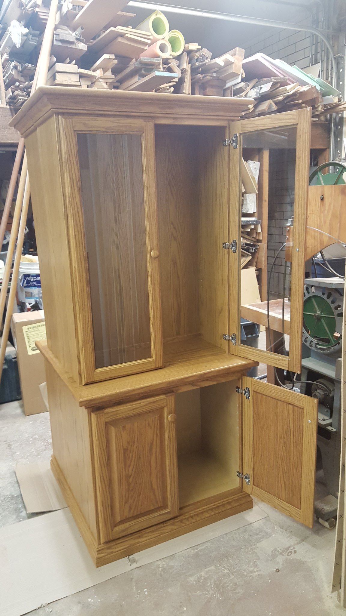 A two-tone wooden cabinet with glass-front doors on the upper section and solid doors on the lower section, situated in a workshop. The cabinet has four doors with knobs, and is unfinished inside. The background shows various woodworking tools and ma