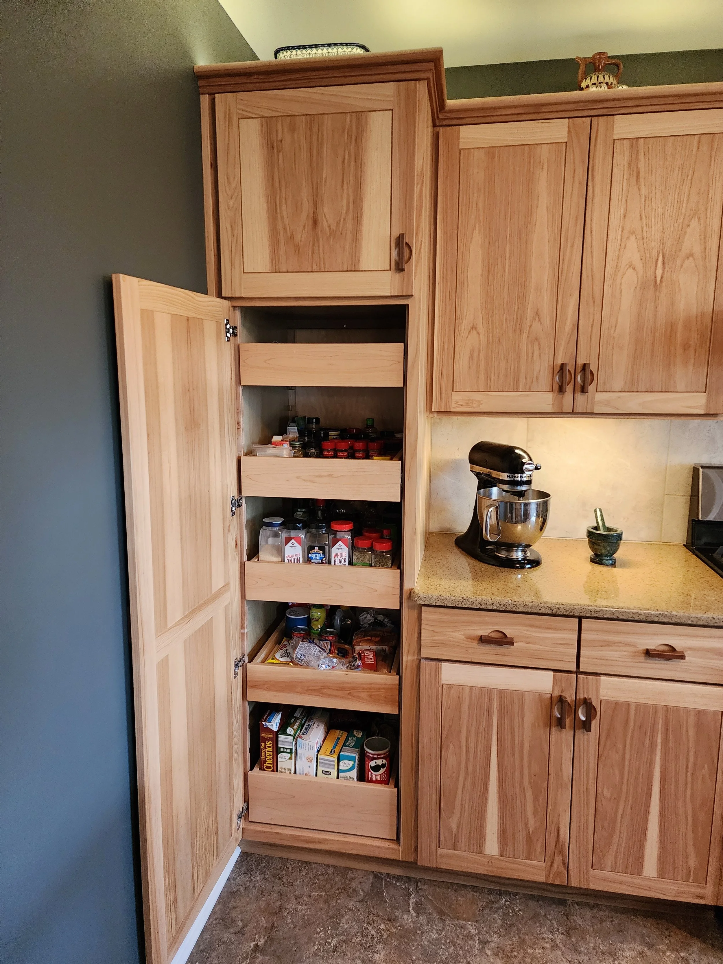 Open kitchen pantry with spices and food items beside a stand mixer on a beige countertop and wooden cabinets.