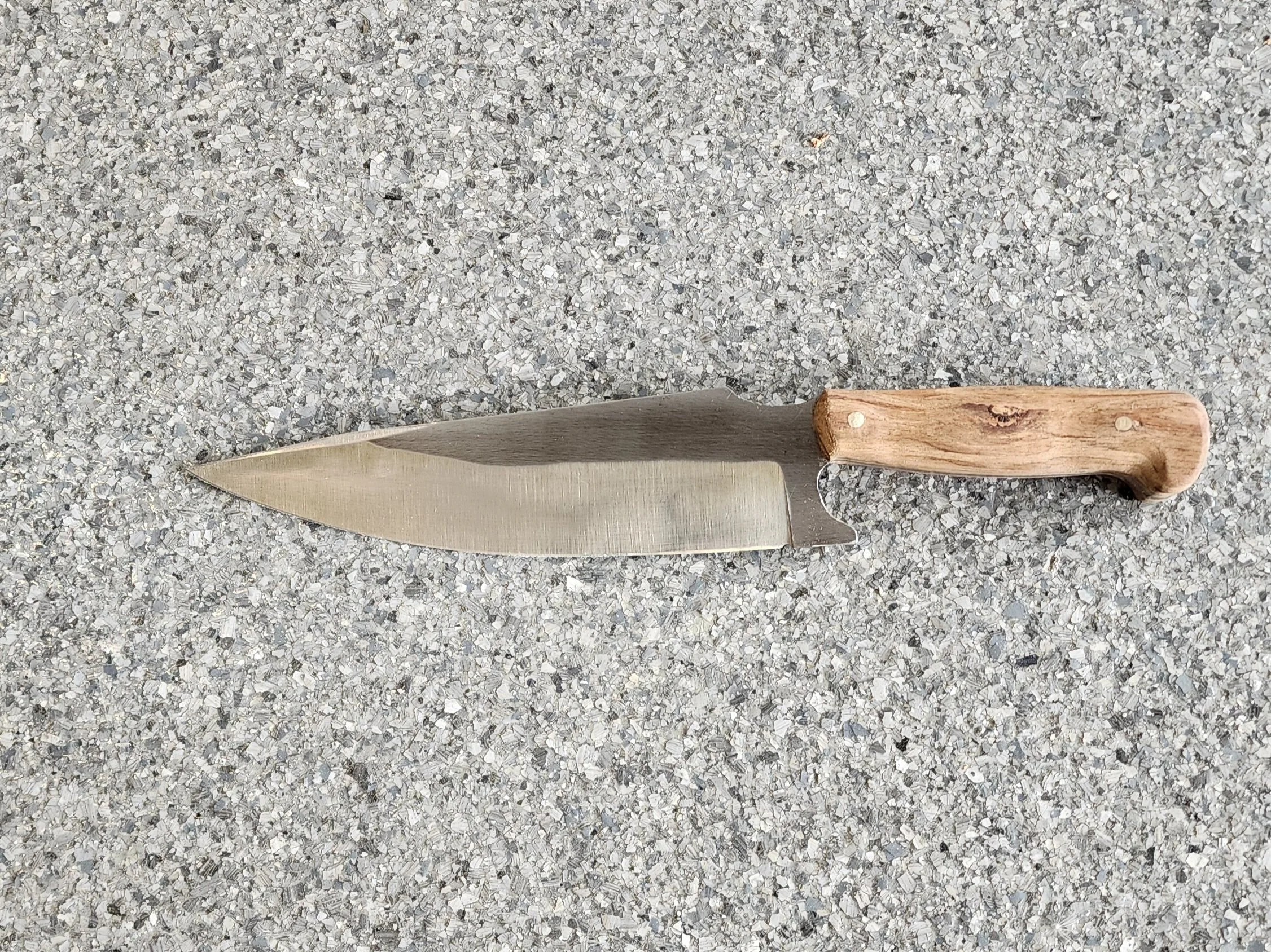 A large kitchen knife with a wooden handle and a broad, stainless steel blade resting on a textured gray surface.