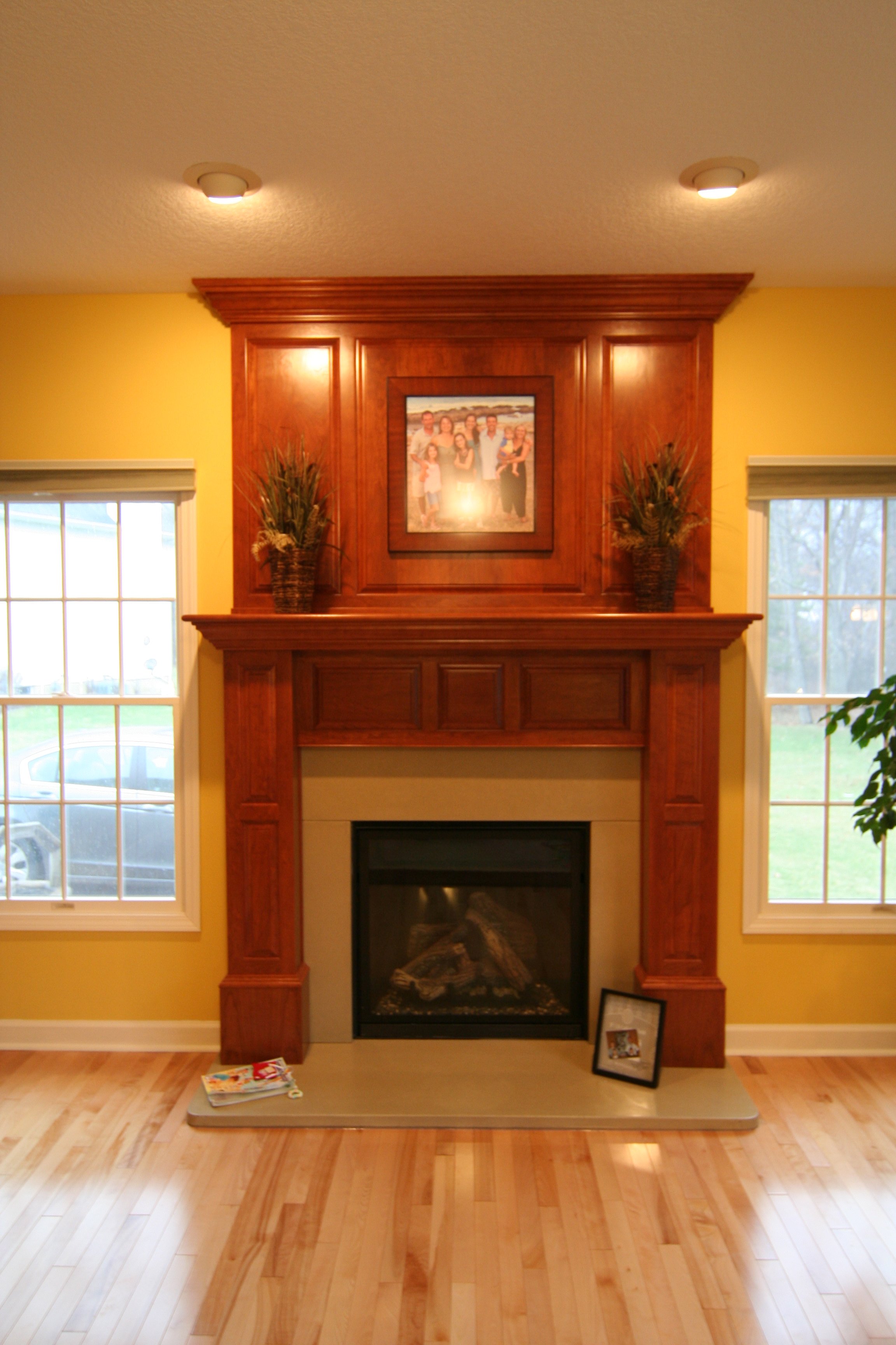 A living room with a yellow wall and a wooden fireplace mantel. The mantel has two potted plants, one on each side, and a framed family photo above it. There are two windows on either side of the fireplace with blinds, and a framed photo and some mag