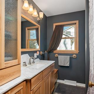 Bathroom with a wooden vanity, white countertop, large mirror, four-light vanity fixture, two small windows with brown curtains, towel bar, and dark gray walls.