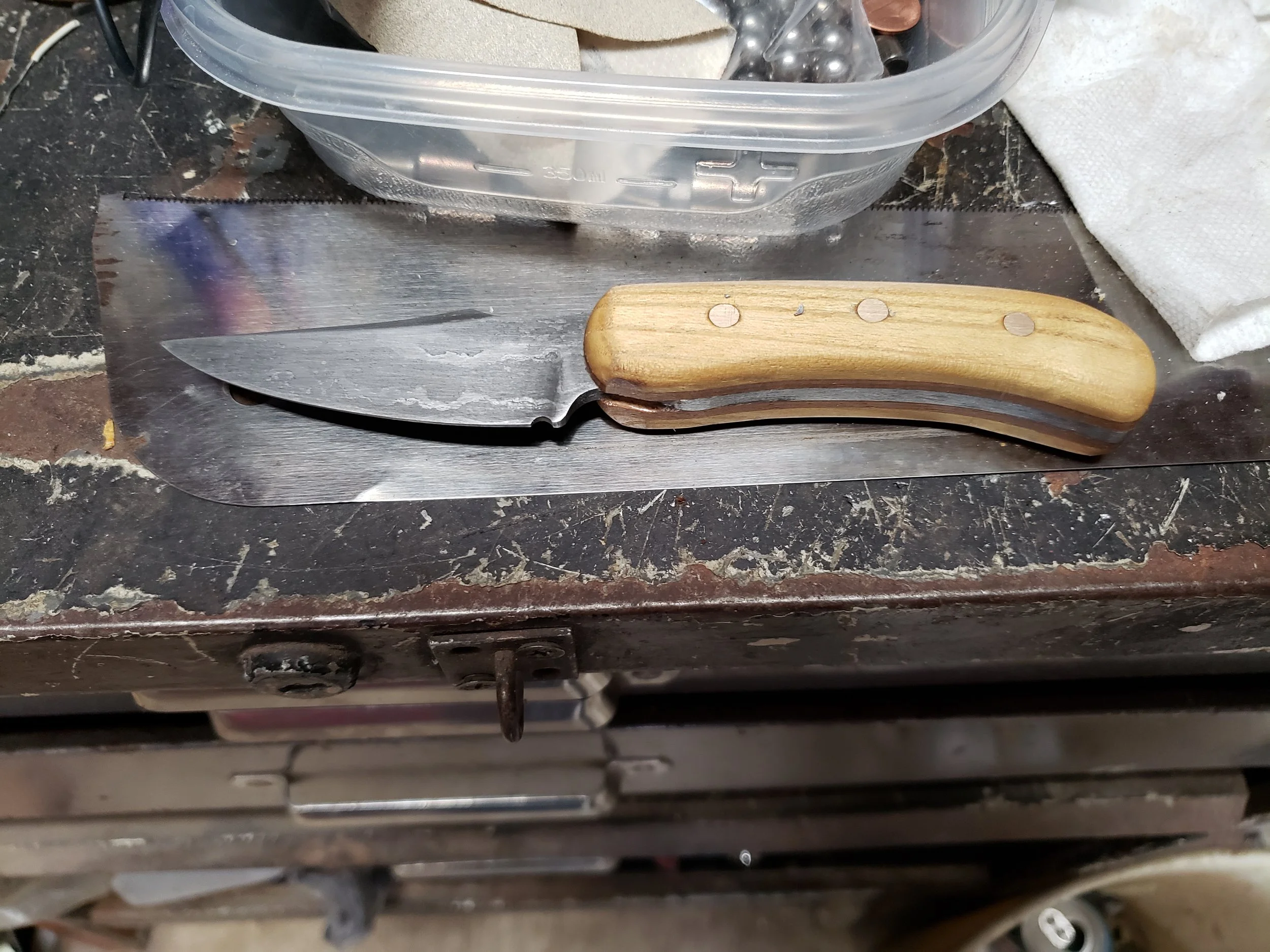 A kitchen knife with a wooden handle resting on a metal sheet, with a plastic container and paper towel nearby.