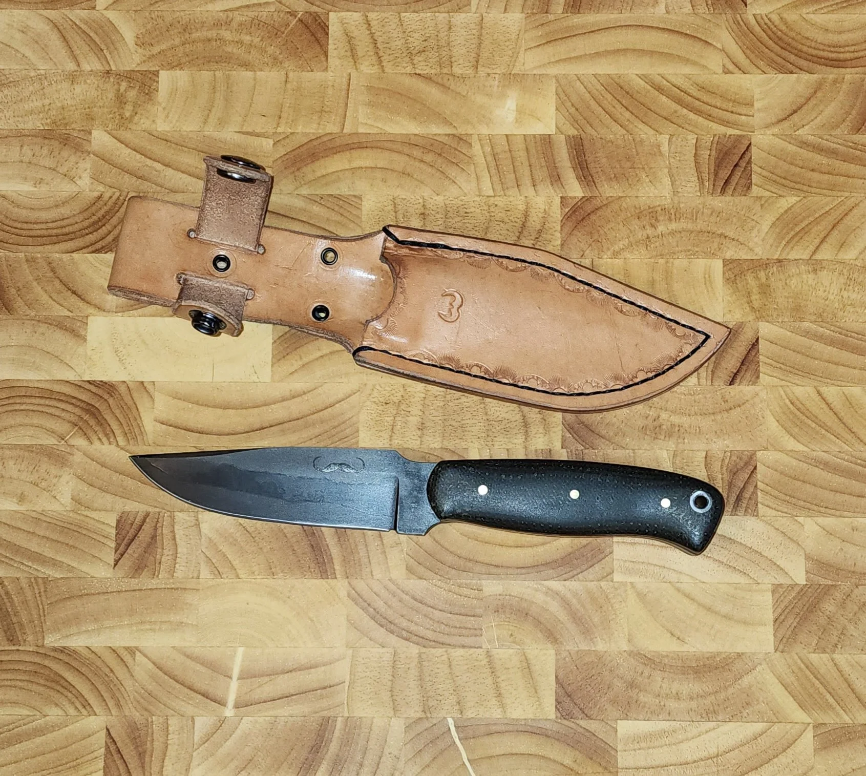 A fixed-blade knife with a black handle and a curved blade, positioned beneath a leather sheath on a wooden surface.