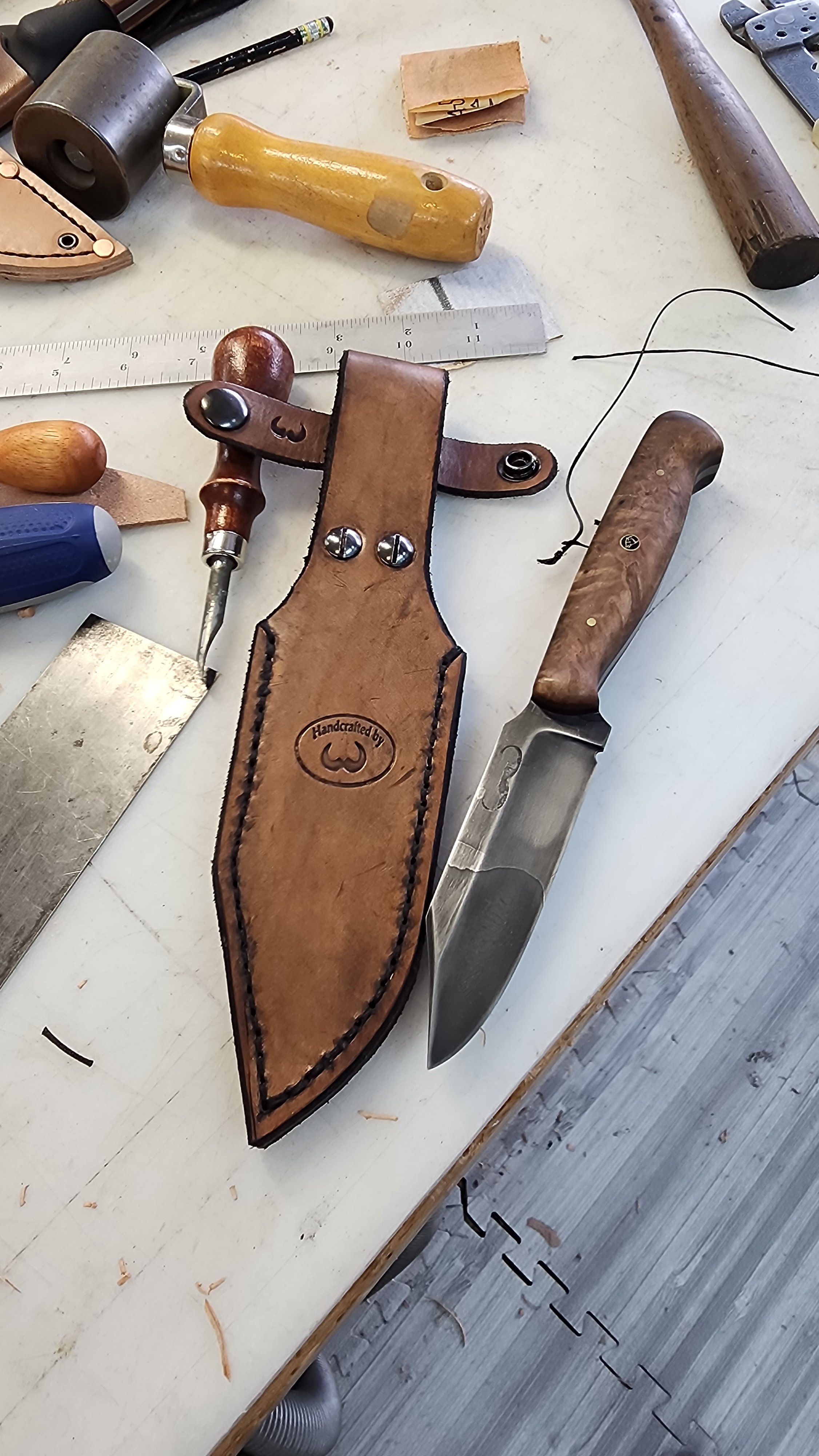 Leatherworking tools and a partially sheathed fixed-blade knife on a work table.