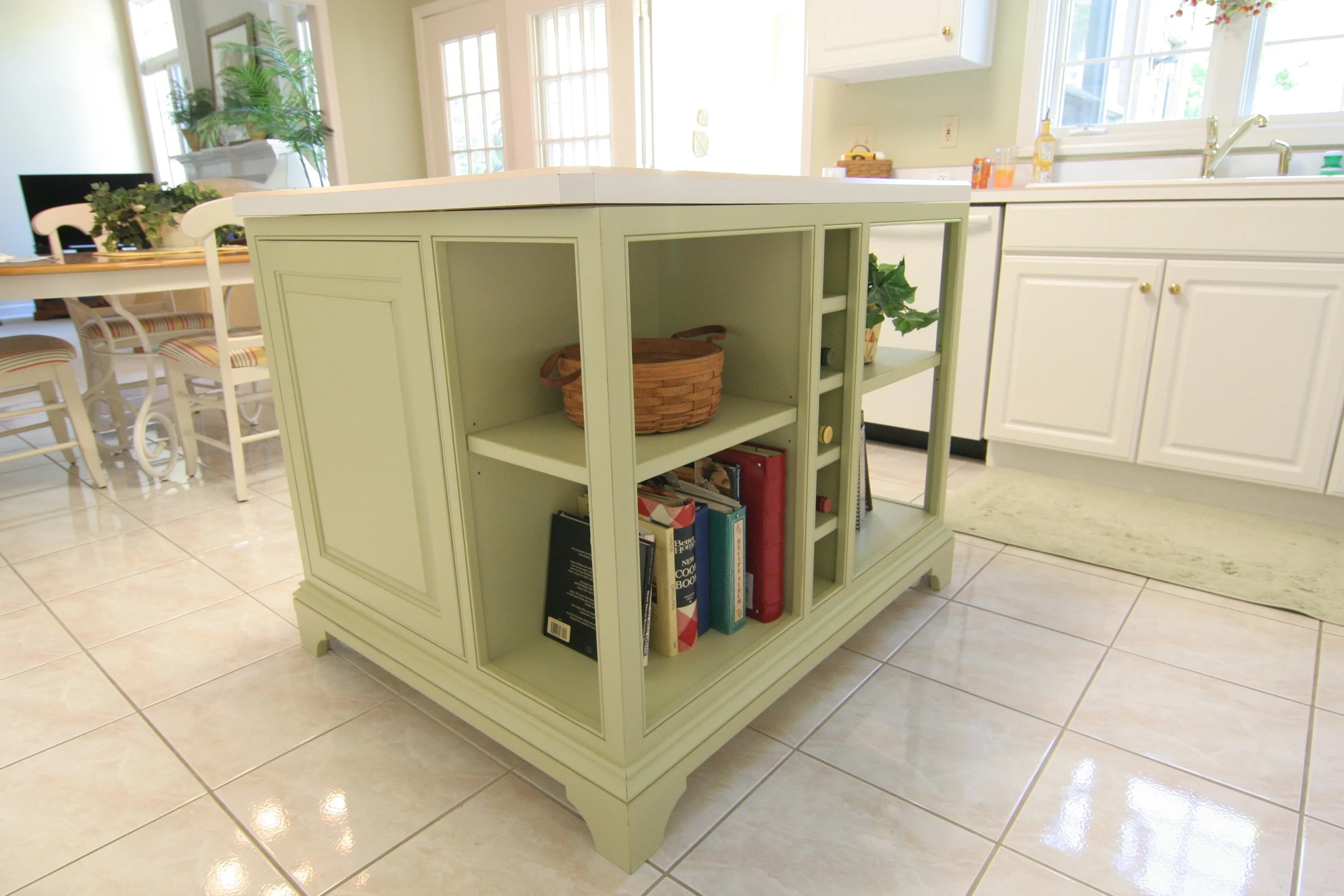 A light green kitchen island with open shelving, books, and a basket, situated in a bright kitchen with white cabinets and large windows.
