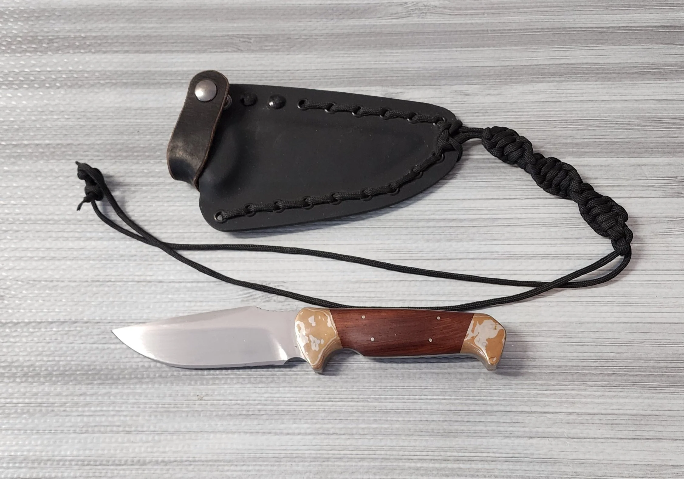 A small fixed-blade pocket knife with a wooden and beige handle, and a black leather sheath with a.paracord lanyard, placed on a light wooden surface.