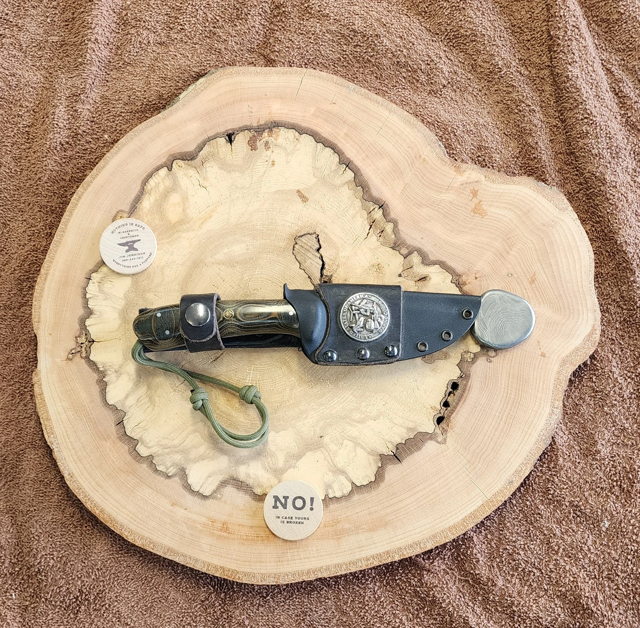 A pocket knife with a black handle and silver blade resting on a circular wooden slab, with two small circular labels on the slab reading 'NOTHING IS SAFE' and 'NO!'.