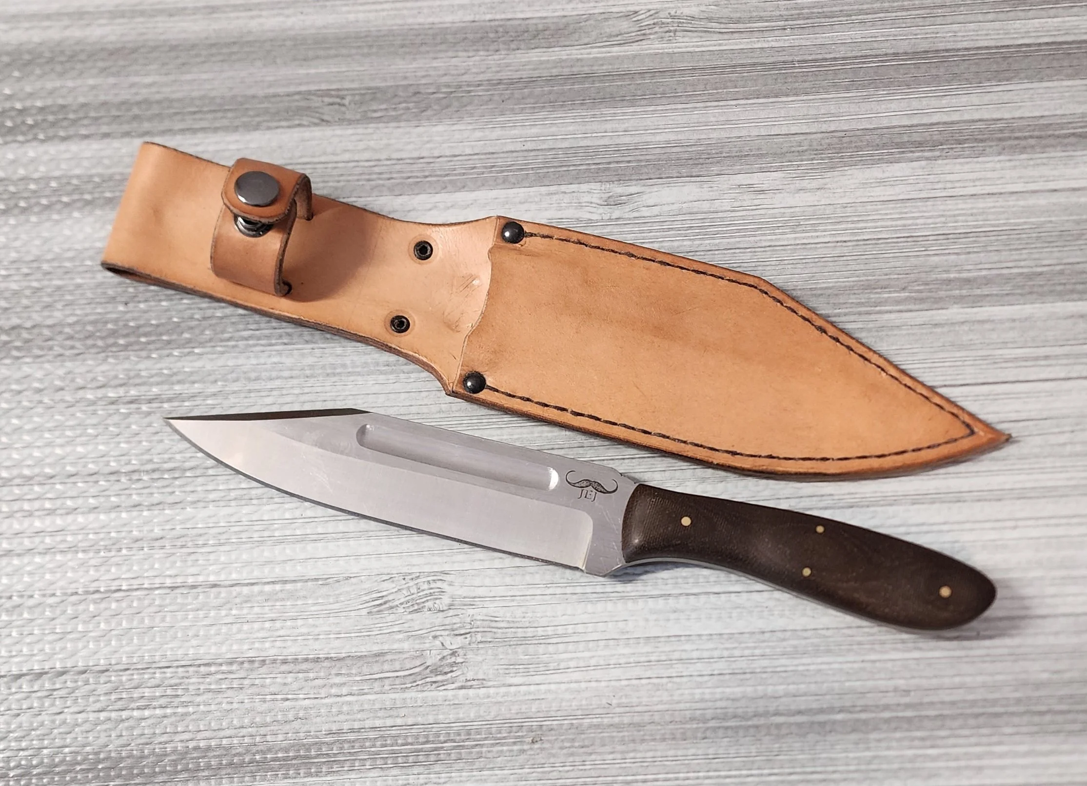 A kitchen knife with a dark handle and logo, along with a tan leather sheath, placed on a light gray wooden surface.