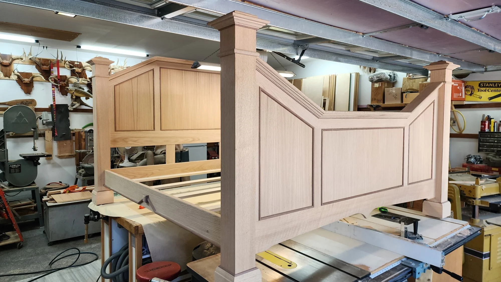 Wooden bed frame being constructed in a woodworking shop, with various tools and wood pieces around.