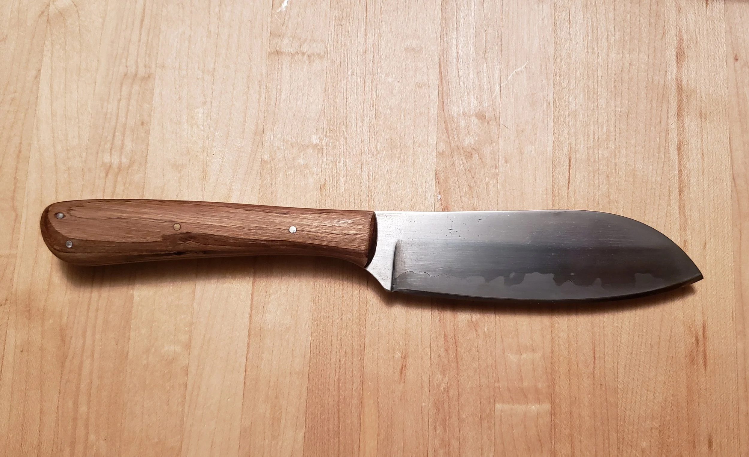 A kitchen knife with a wooden handle and a dark, slightly curved blade is placed on a light wooden surface.