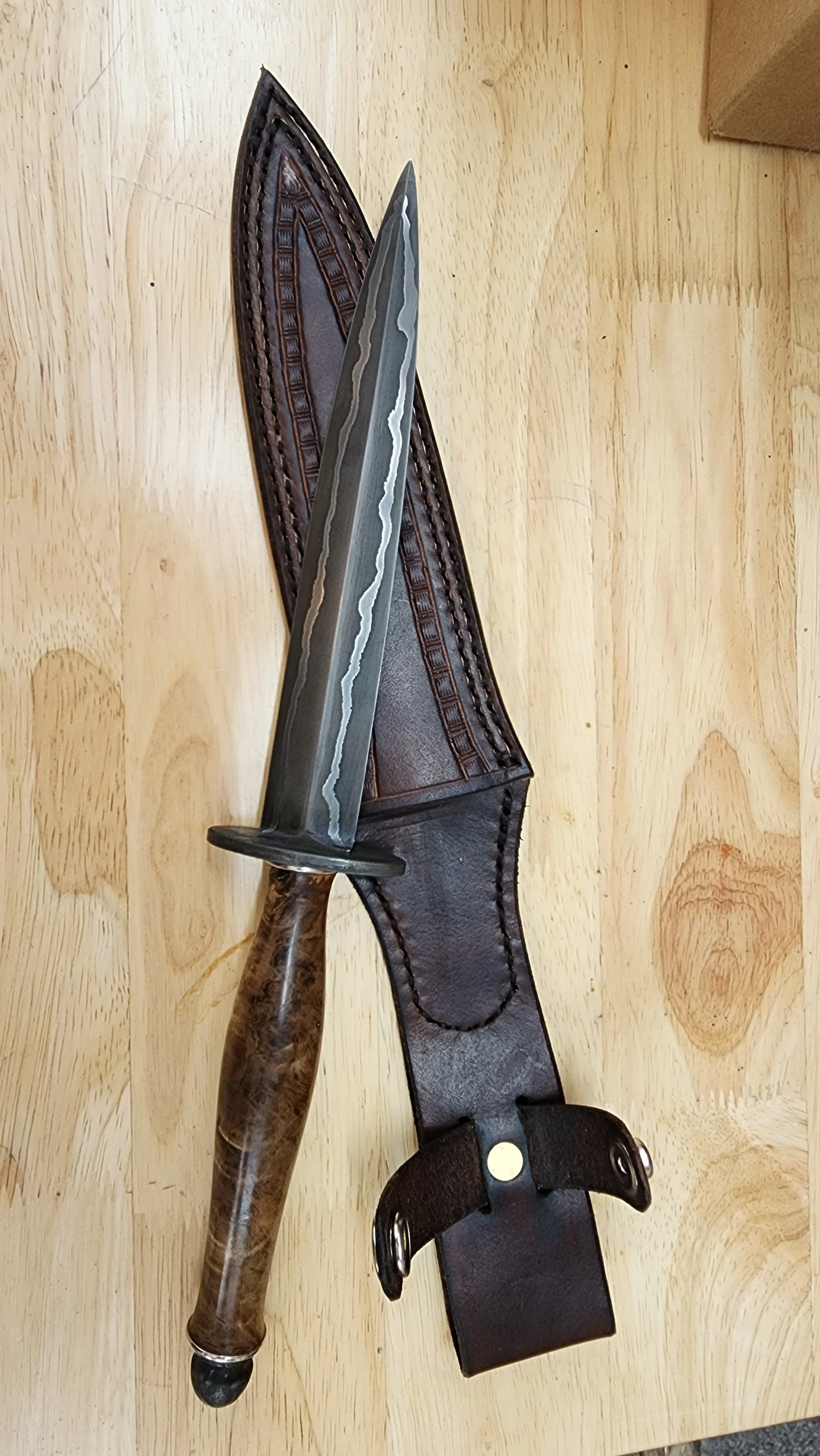 A large knife with a wooden handle lying on a leather sheath on a wooden surface.