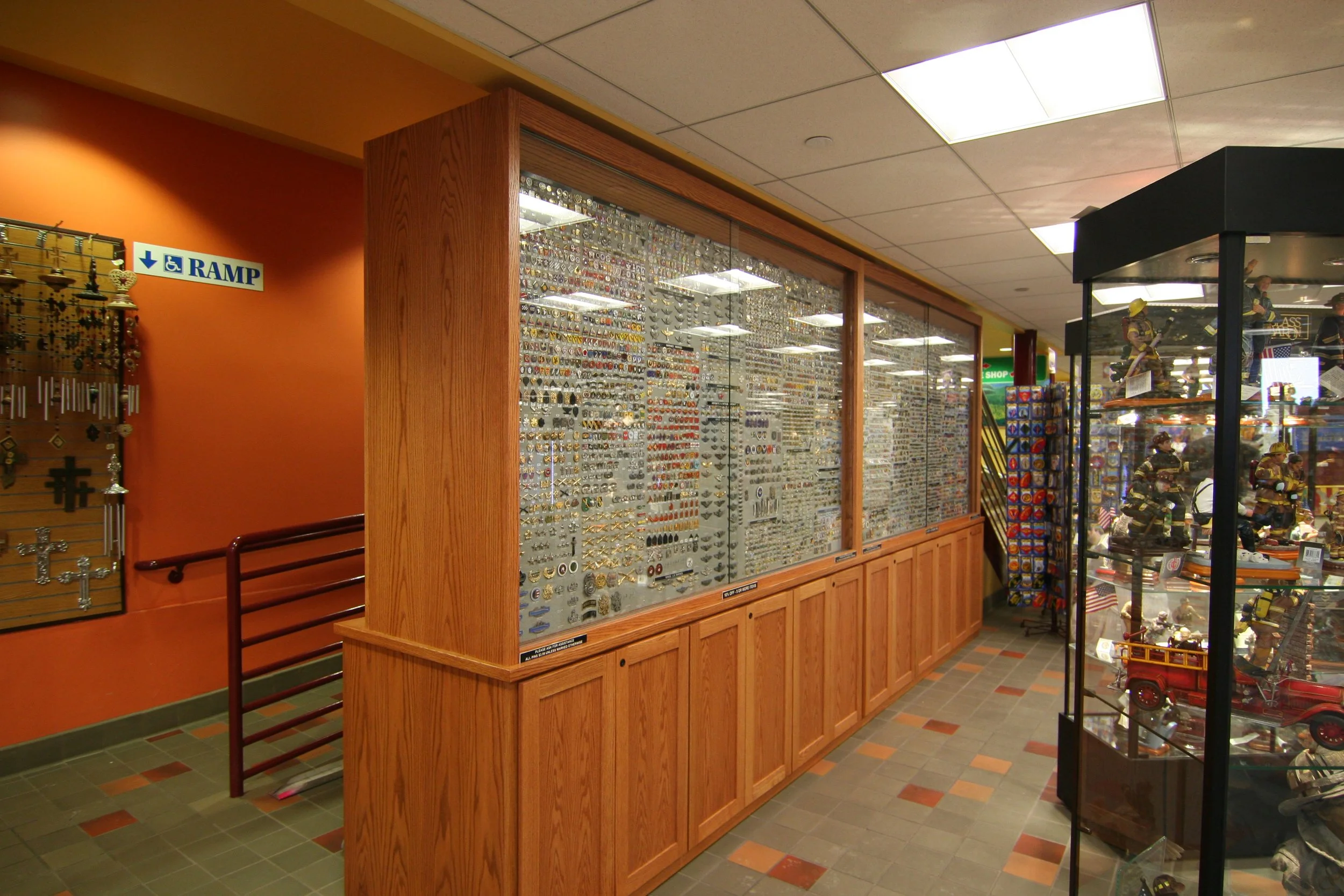 Display case filled with various pins and patches inside a retail store. Firefighter mannequins dressed in gear are visible on the right. The store has a colorful tiled floor and wooden framing around the display case.