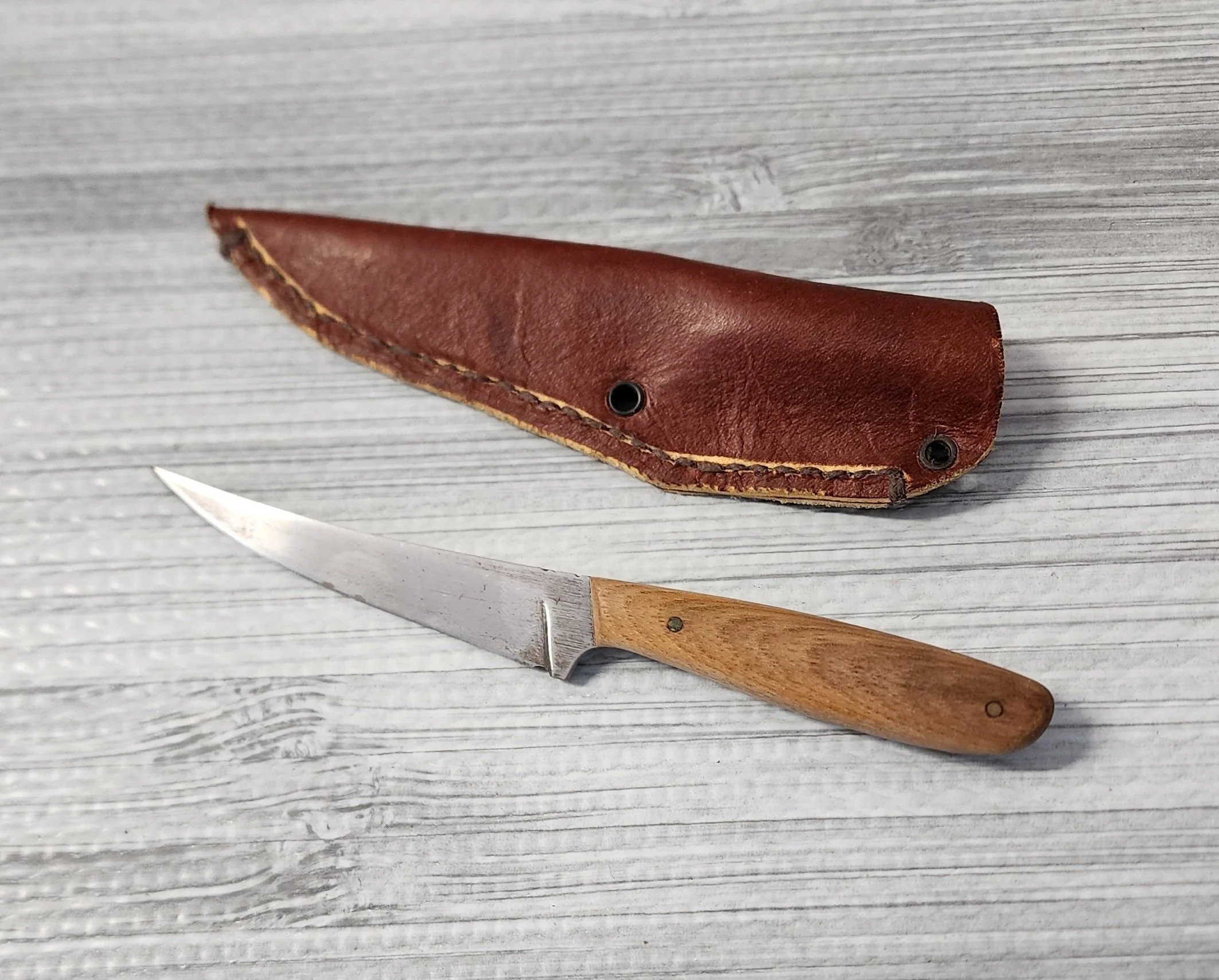 A small pocket knife with a wooden handle and a metal blade, alongside a brown leather case, on a light wooden surface.