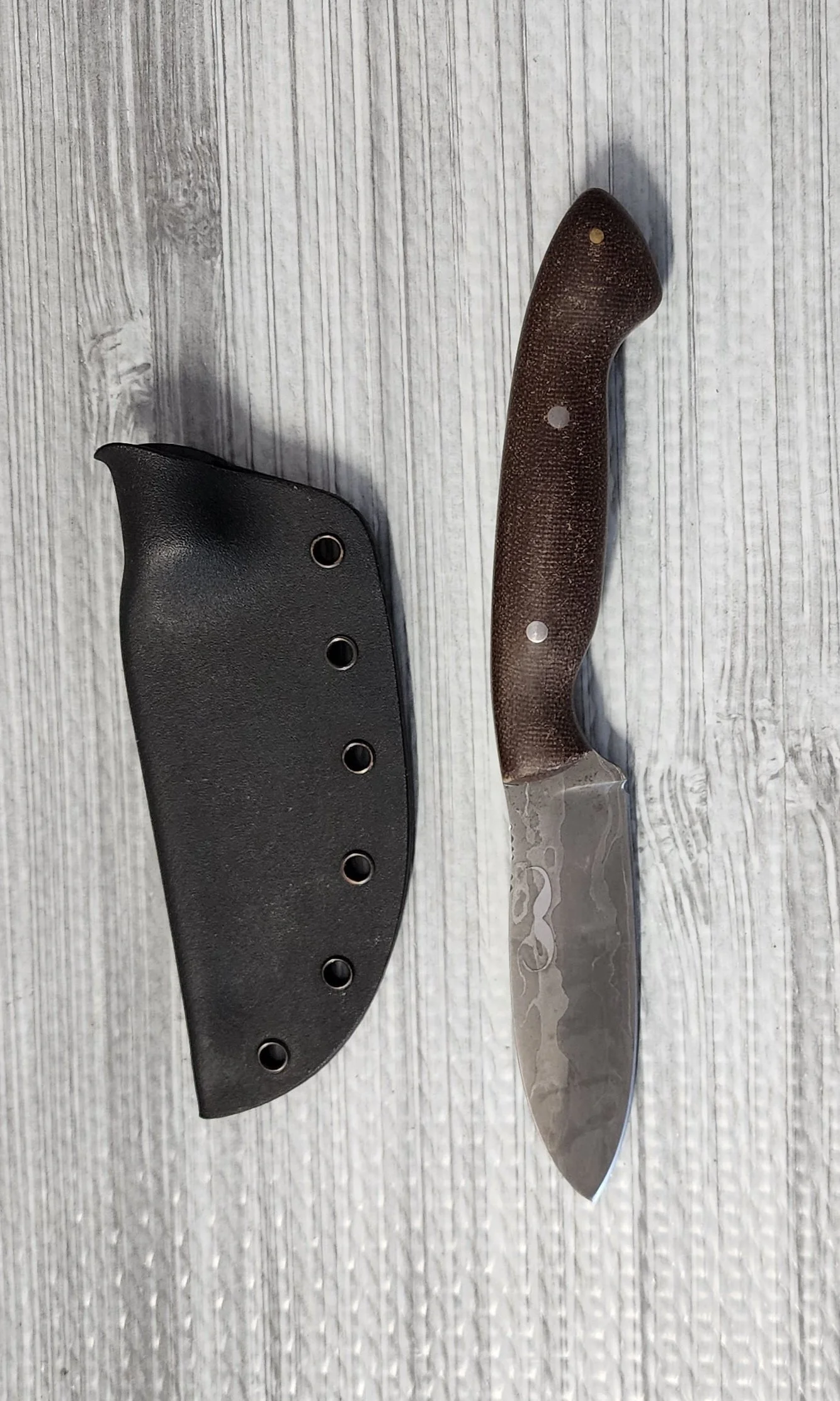 A fixed-blade knife with a brown textured handle and a patterned metal blade next to a black knife sheath on a white wooden surface.