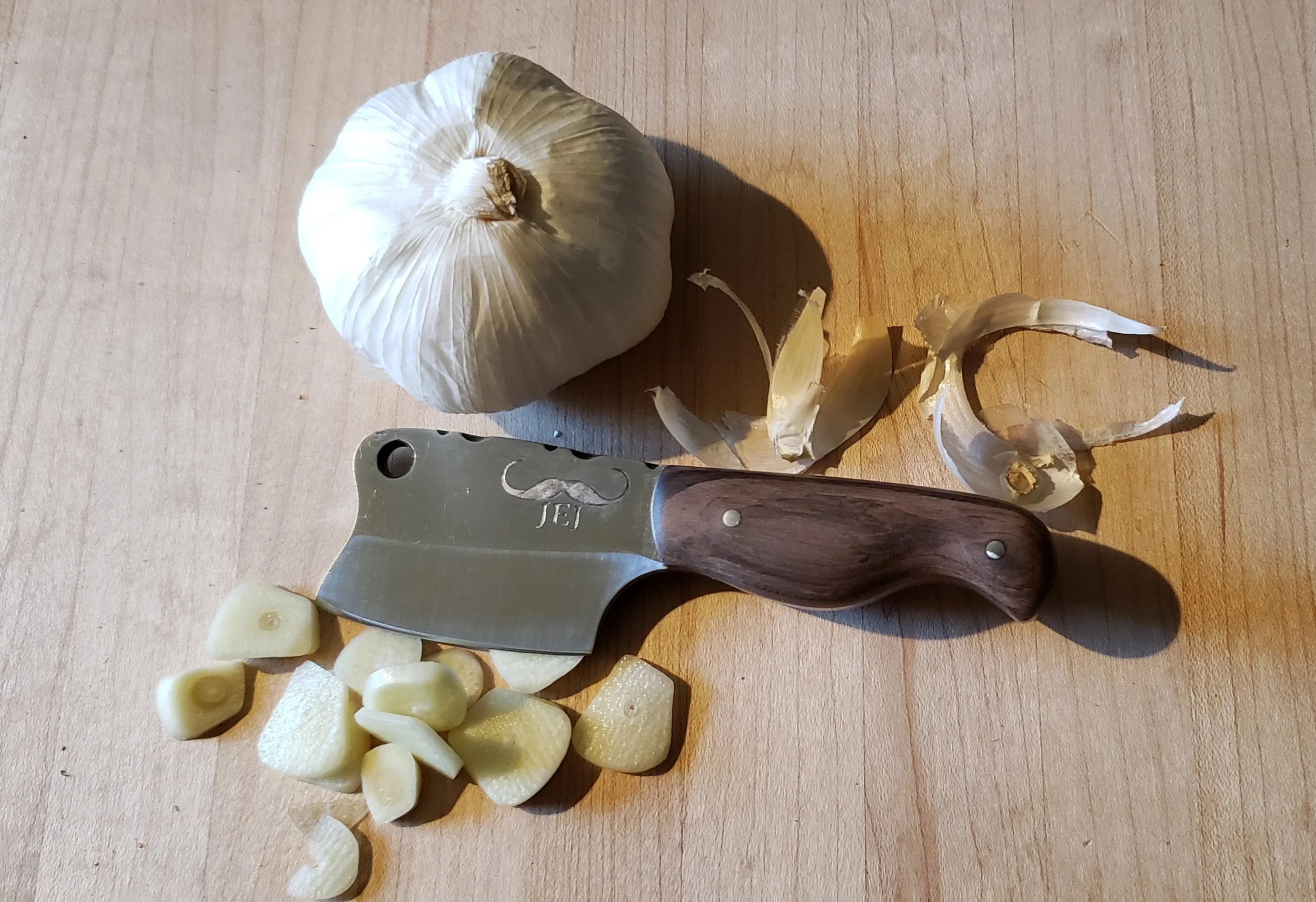 Whole garlic bulb, garlic cloves, garlic peel, and a garlic knife on a wooden cutting board.