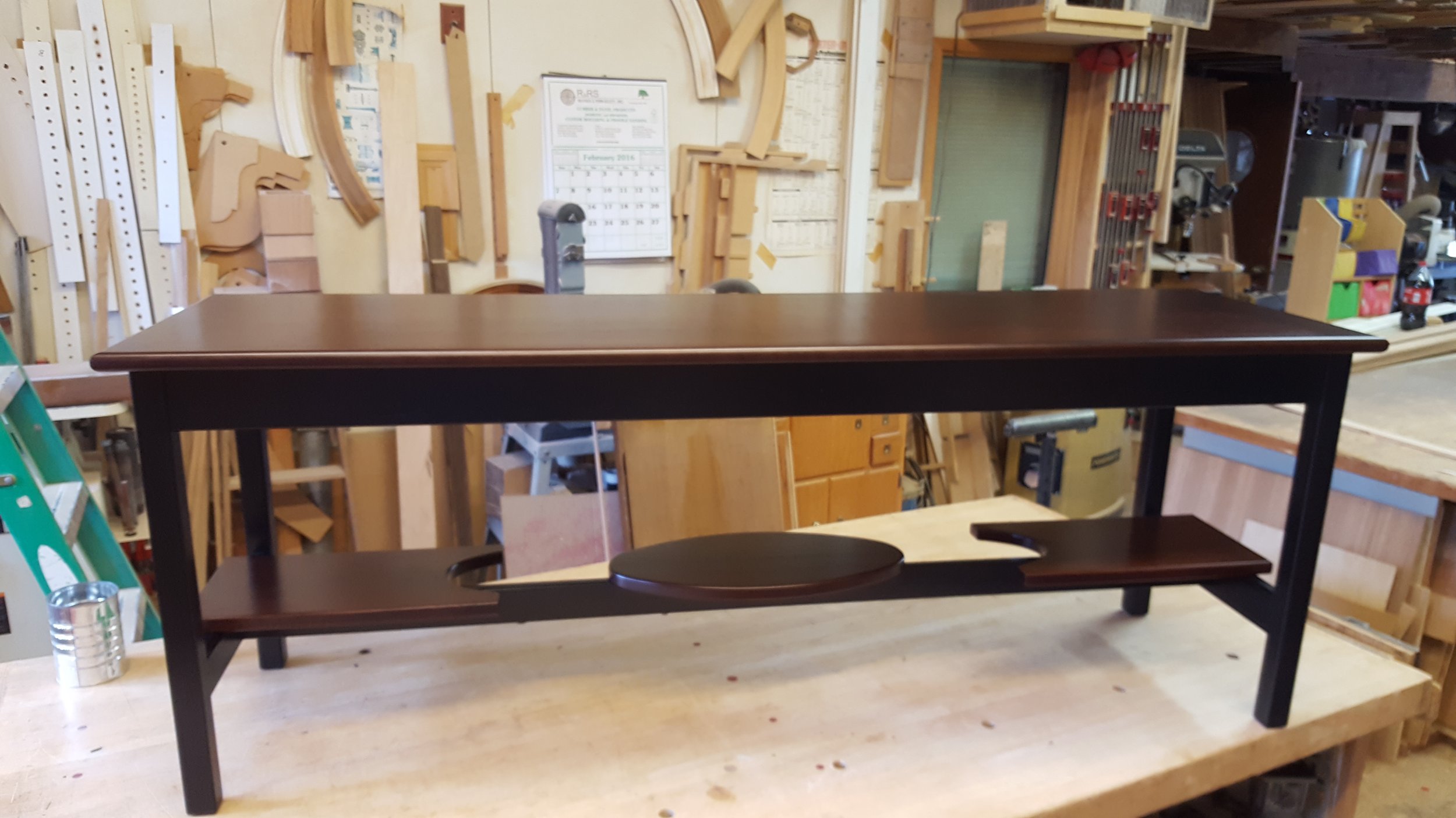 A dark wood dining table with a circular tray and an extension leaf on a wooden workbench in a woodworking shop.