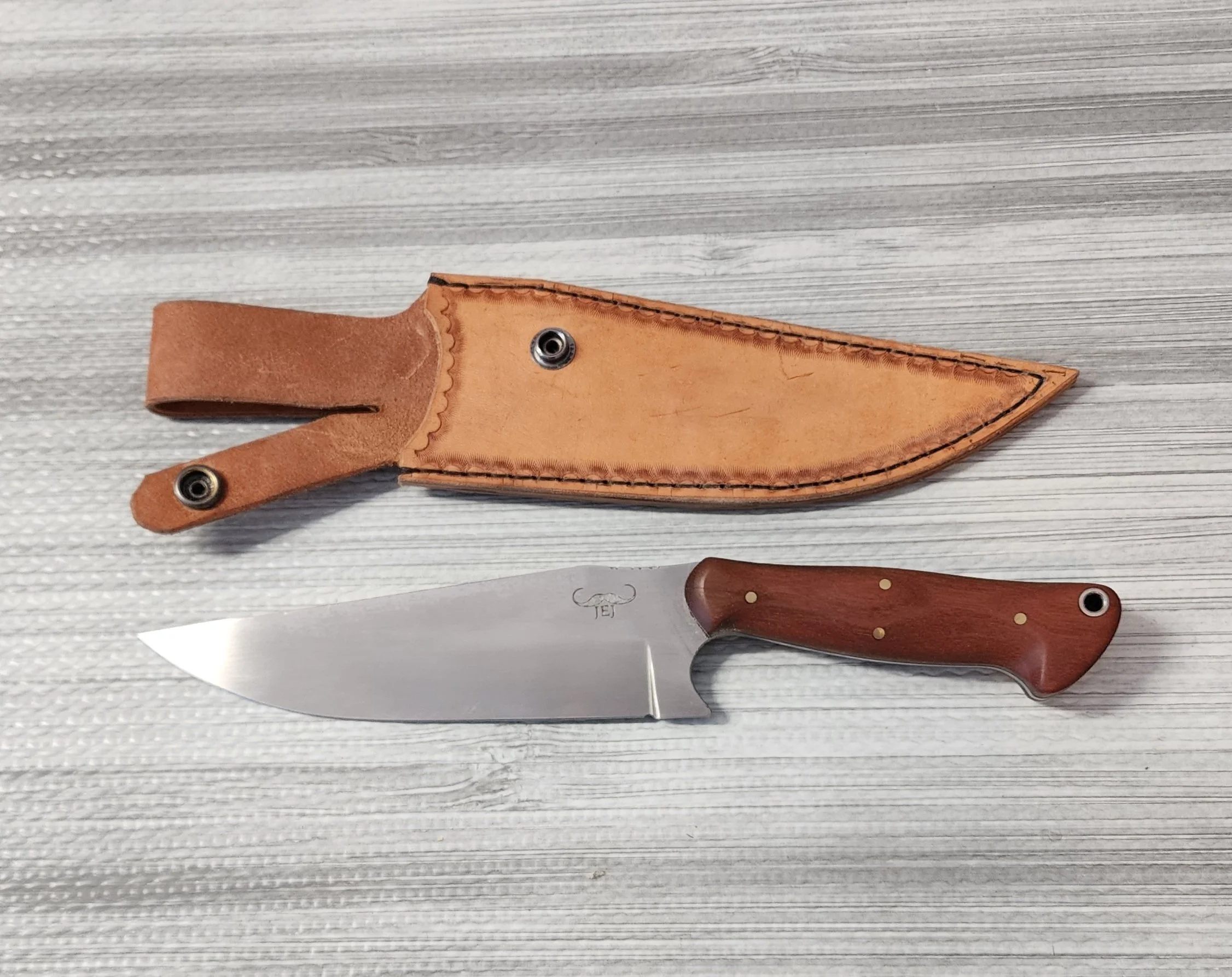 A hunting or utility knife with a wooden handle, a stainless steel blade, and a leather sheath, all placed on a light-colored wooden surface.