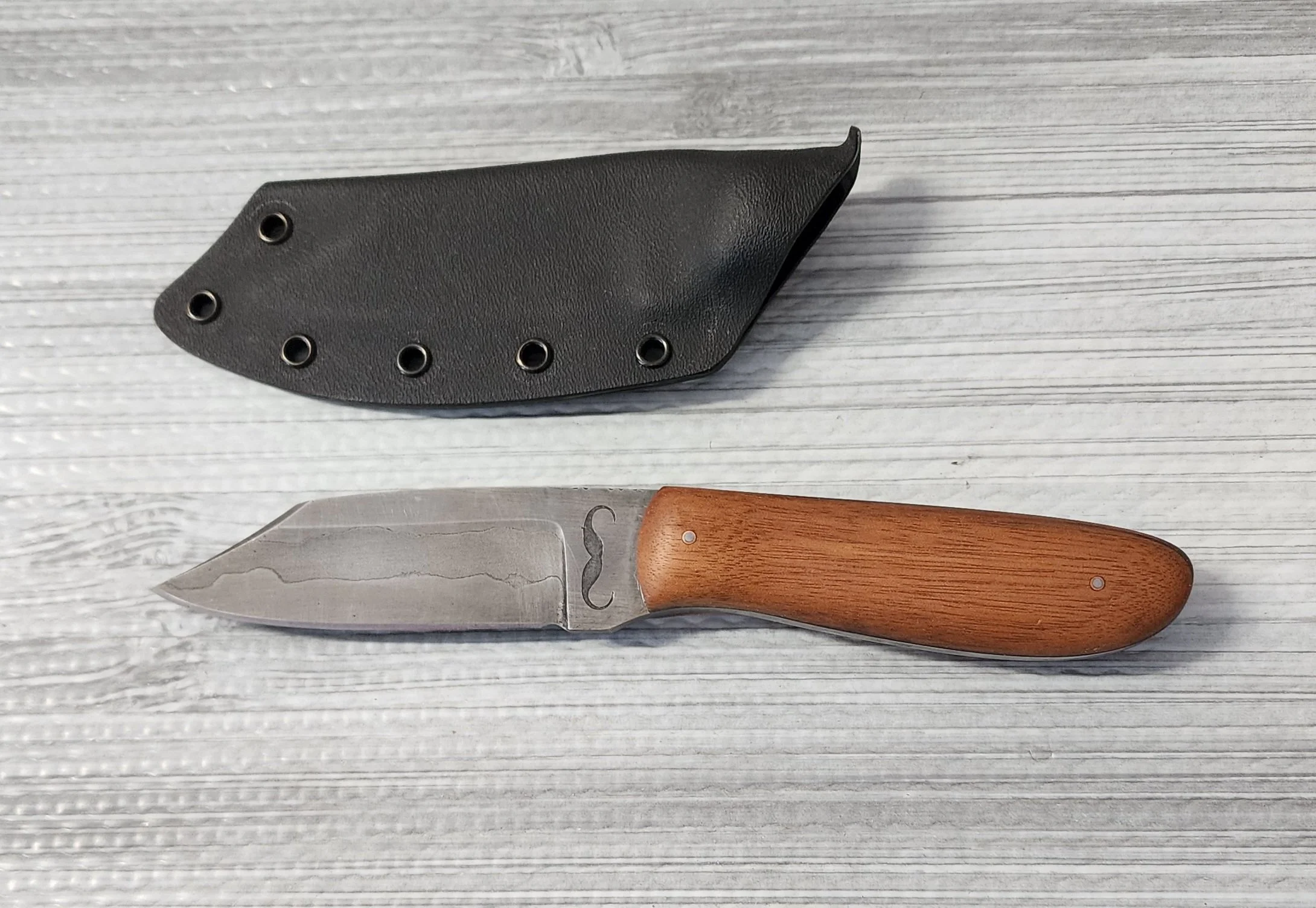 A pocket folding knife with a wooden handle and a stainless steel blade, placed on a light-colored textured surface, alongside a black sheath with multiple rivets.