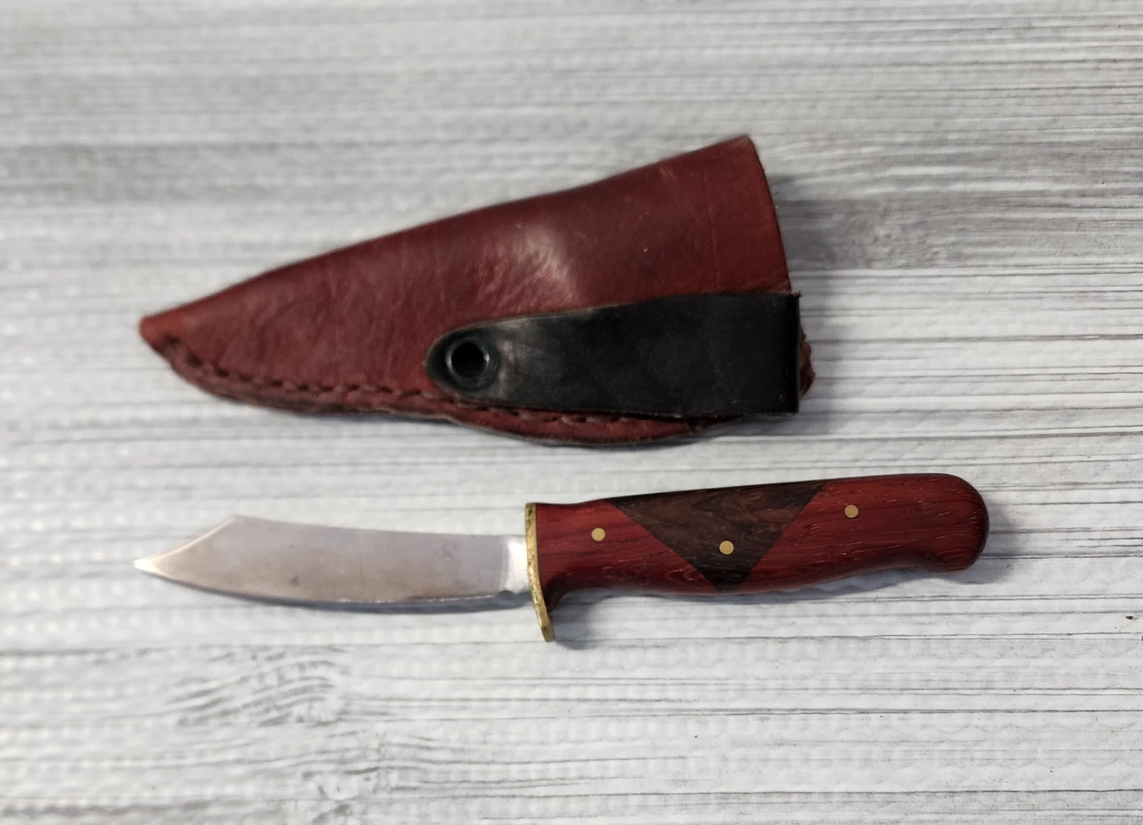 A small folding pocket knife with a red and black wood handle and a slightly worn silver blade, placed next to a red leather sheath on a gray wooden surface.